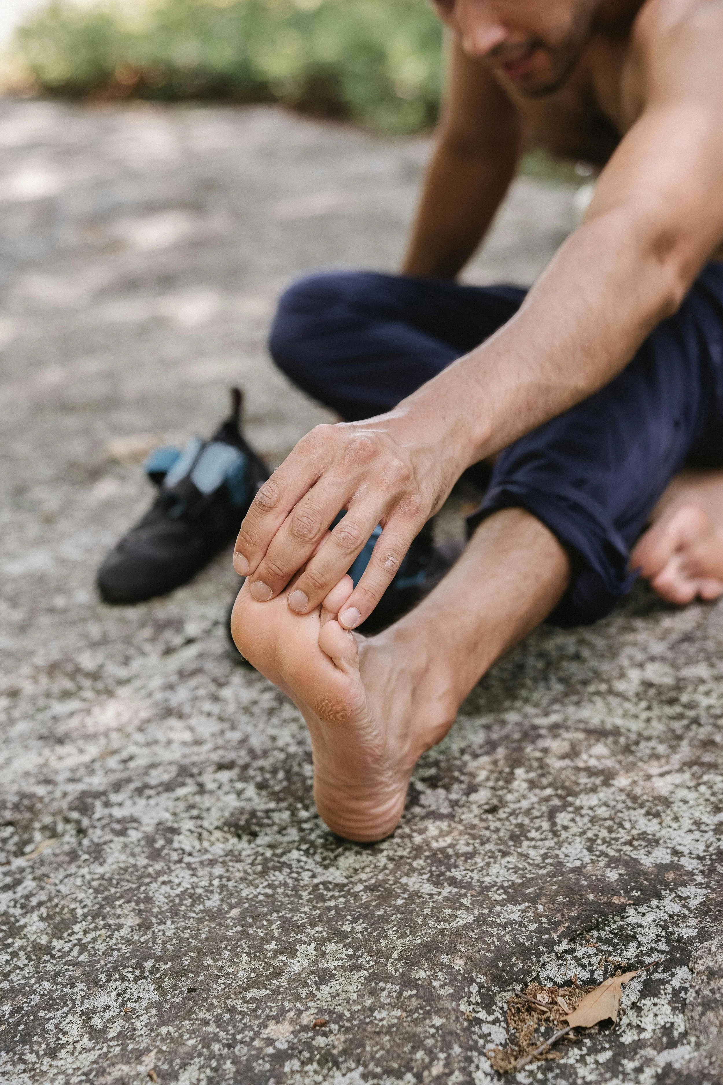 A man sits on a rock outdoors, stretching his legs into a yoga pose while holding his foot with his hand. The scene is natural with trees and fallen leaves visible.