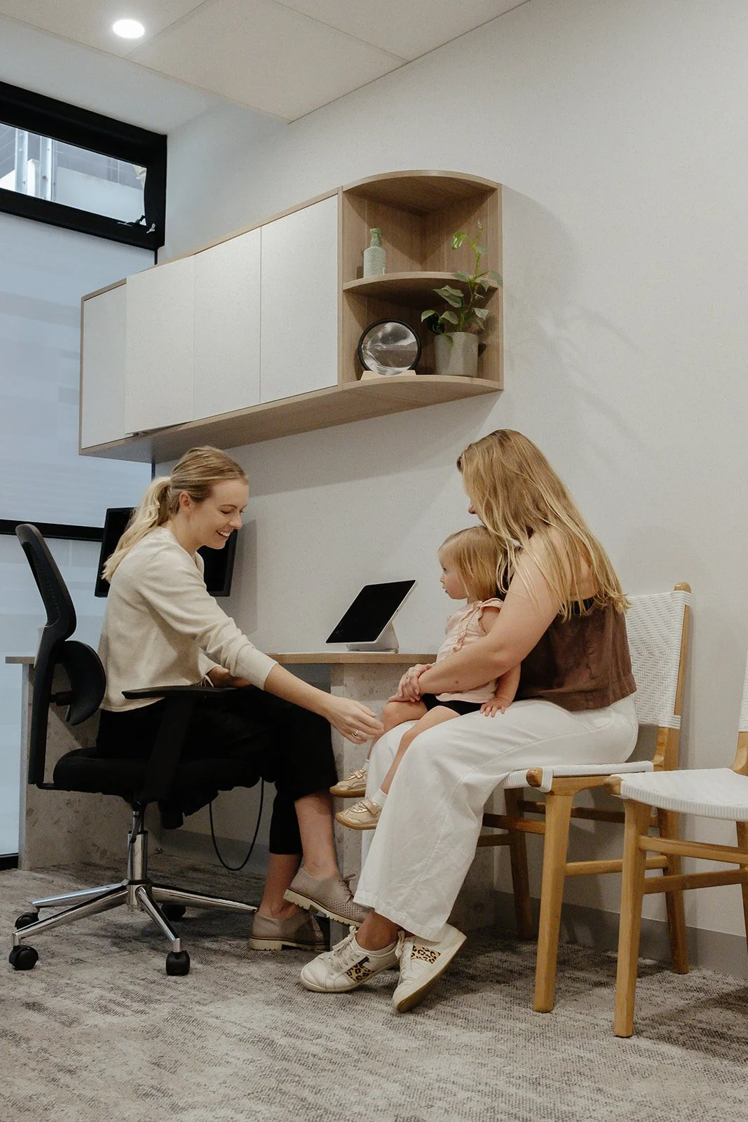 A woman sitting in an office chair is smiling and reaching out to hold the hand of a young girl. The young girl is sitting on a woman's lap, holding hands with her, and looking at the woman in the office chair. There is a desk, a computer, and a wall-mounted cabinet with shelves and plants in the background.