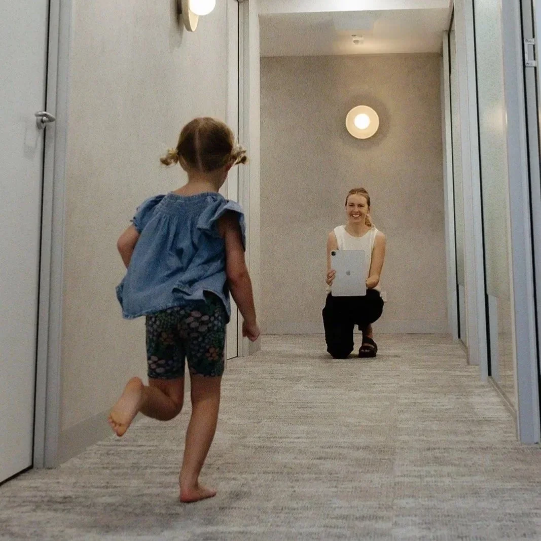 A young girl running towards a woman who is kneeling and taking a picture of her in a hallway with beige walls and carpet, smiling at her.