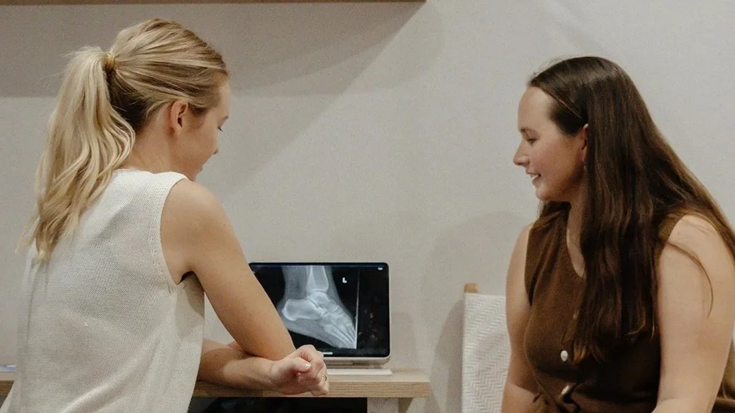 Two women sitting at a table, one showing an X-ray image of a foot on a tablet, and they are smiling.