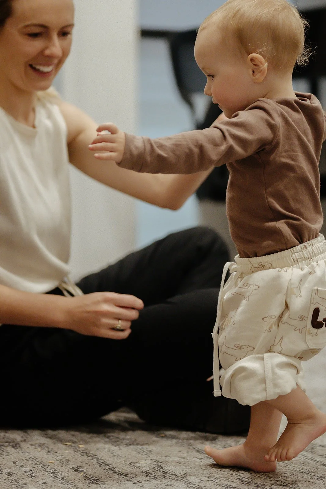 A young child with blond hair standing on a carpeted floor, holding hands with an adult woman, smiling and looking at her. The woman is sitting on the floor wearing a white top and black pants, smiling back at the child.