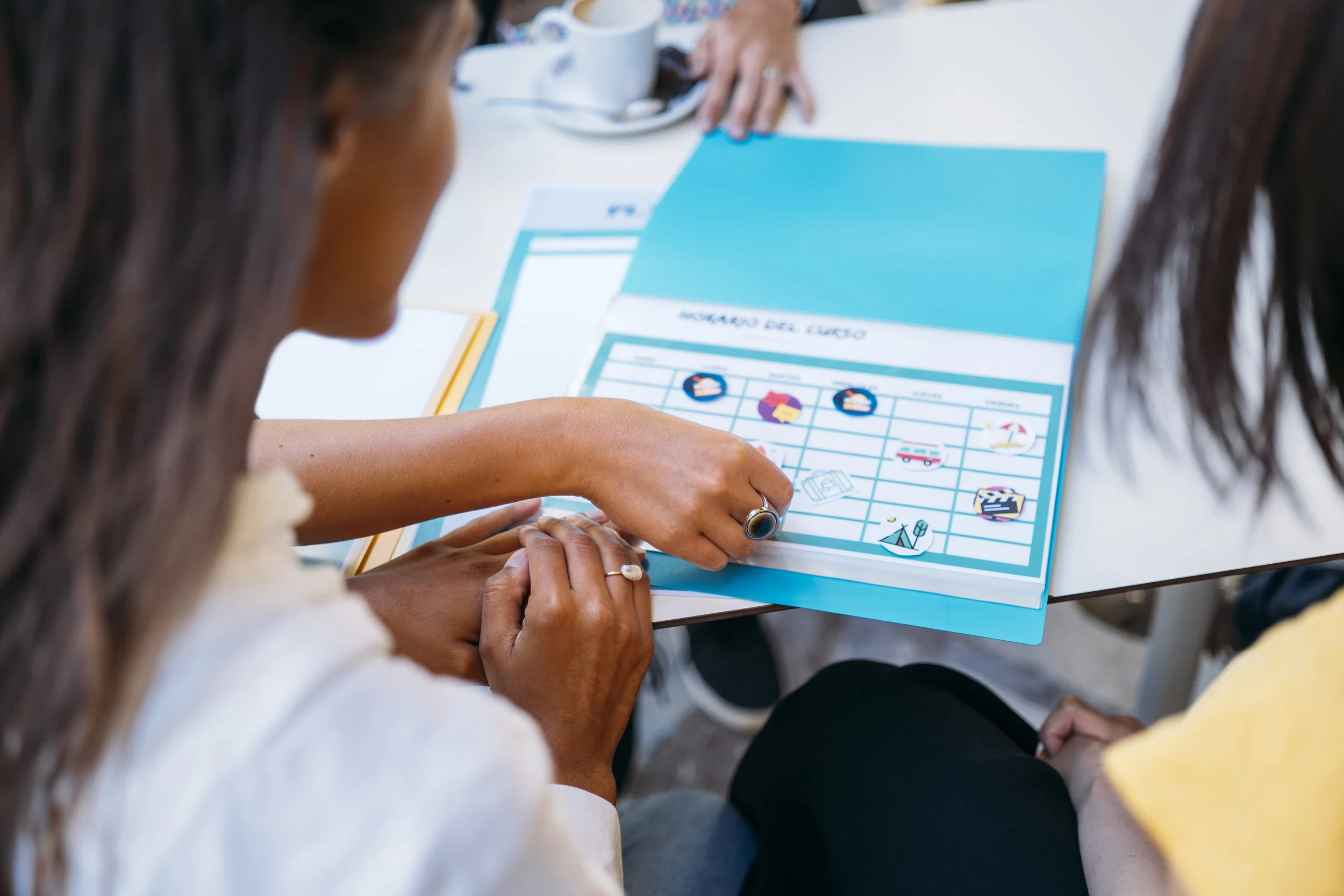 Two women with dark hair sitting at a white table, looking at a blue and white chart or worksheet with travel icons and a grid, engaged in a discussion.