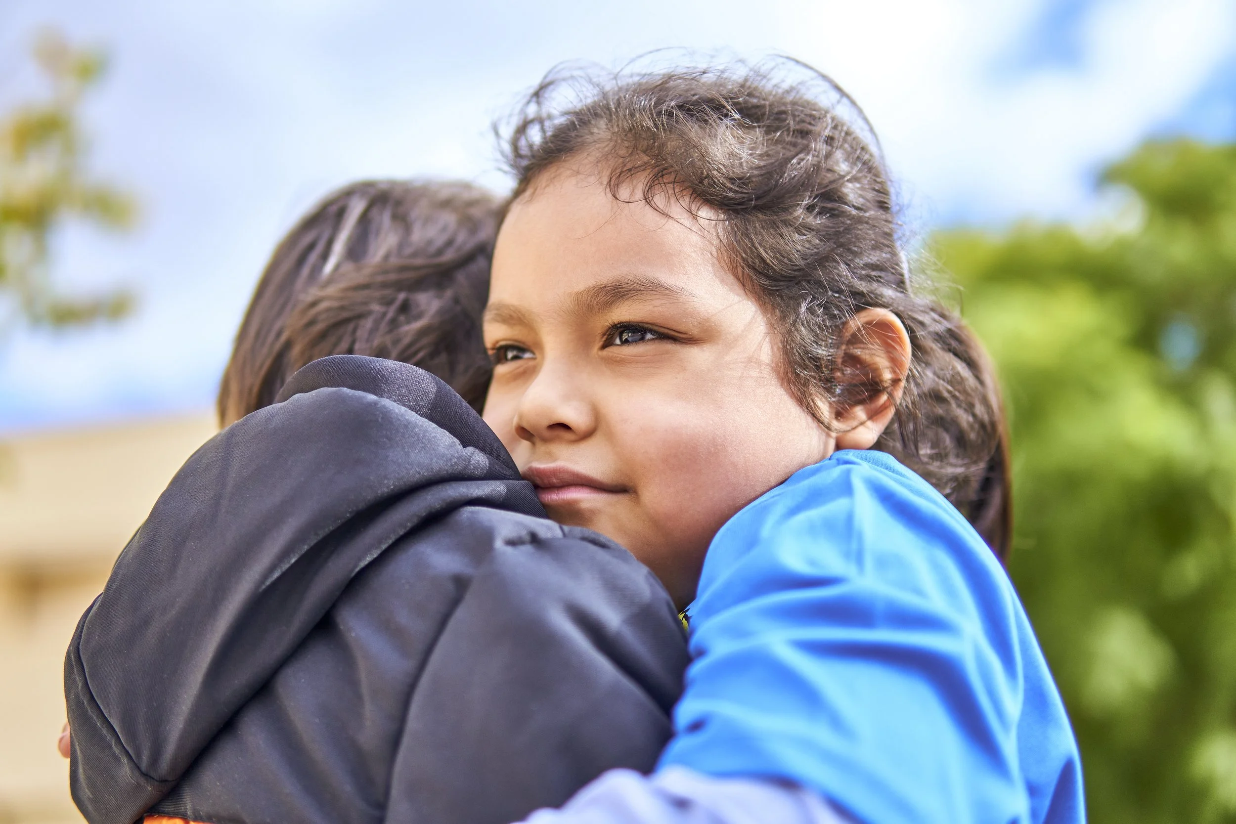 Close-up of two children hugging outdoors on a sunny day, with a background of trees and blue sky.