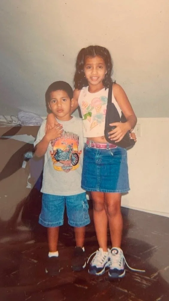 Two children, a girl and a boy, standing indoors on a wooden floor. The girl has curly hair, is wearing a sleeveless crop top with colorful ice cream designs, a denim skirt, and sneakers. The boy has short hair, is wearing a gray T-shirt with a motorcycle graphic, denim shorts, and sneakers. They are posing together, with the girl holding a black bag.