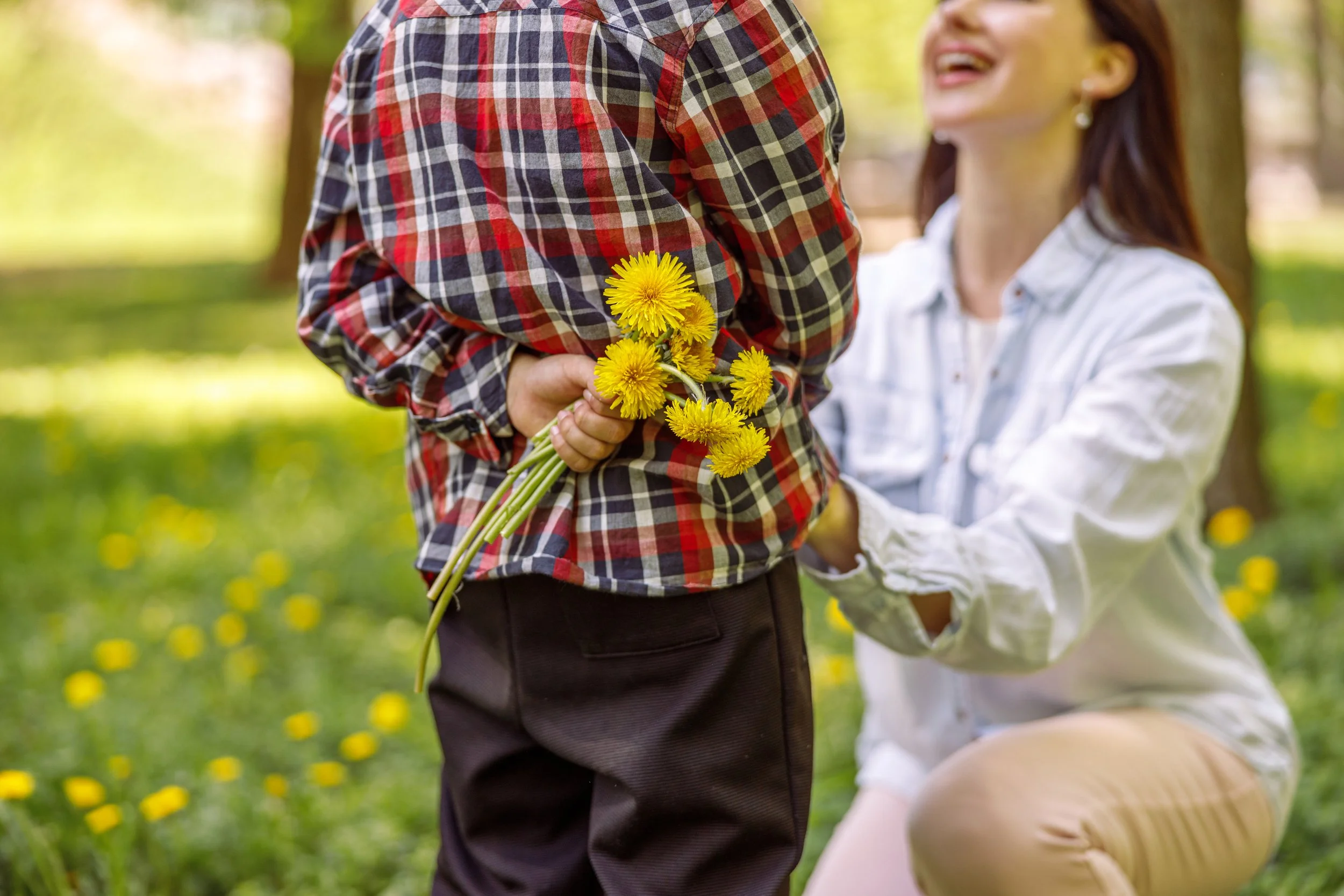 A person holding a bunch of yellow flowers while smiling at a child in an outdoor park setting with green trees and grass in the background.