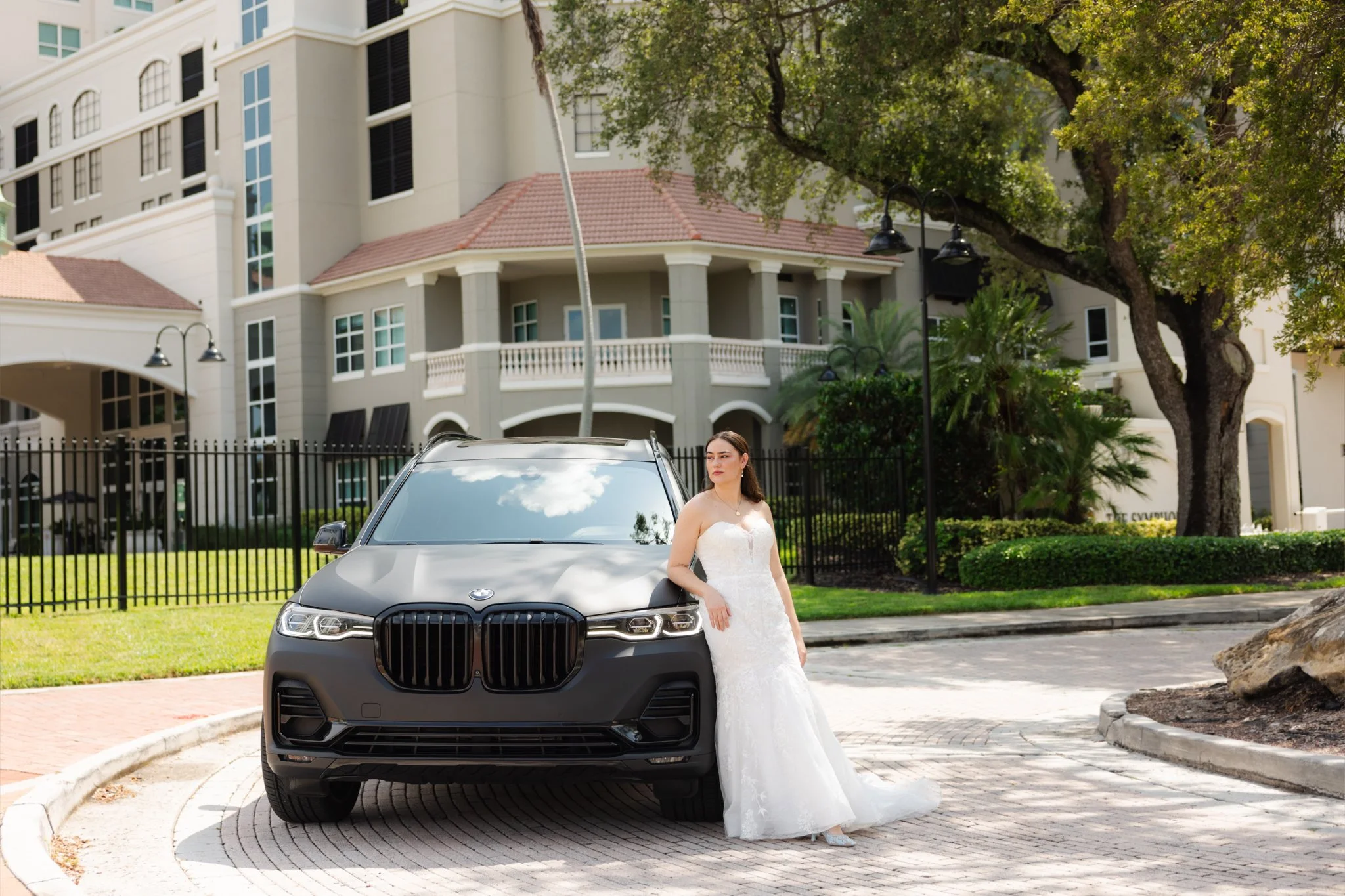 A bride in a white wedding dress leaning against a black luxury car parked in front of a residential building with a fenced yard and large green trees.