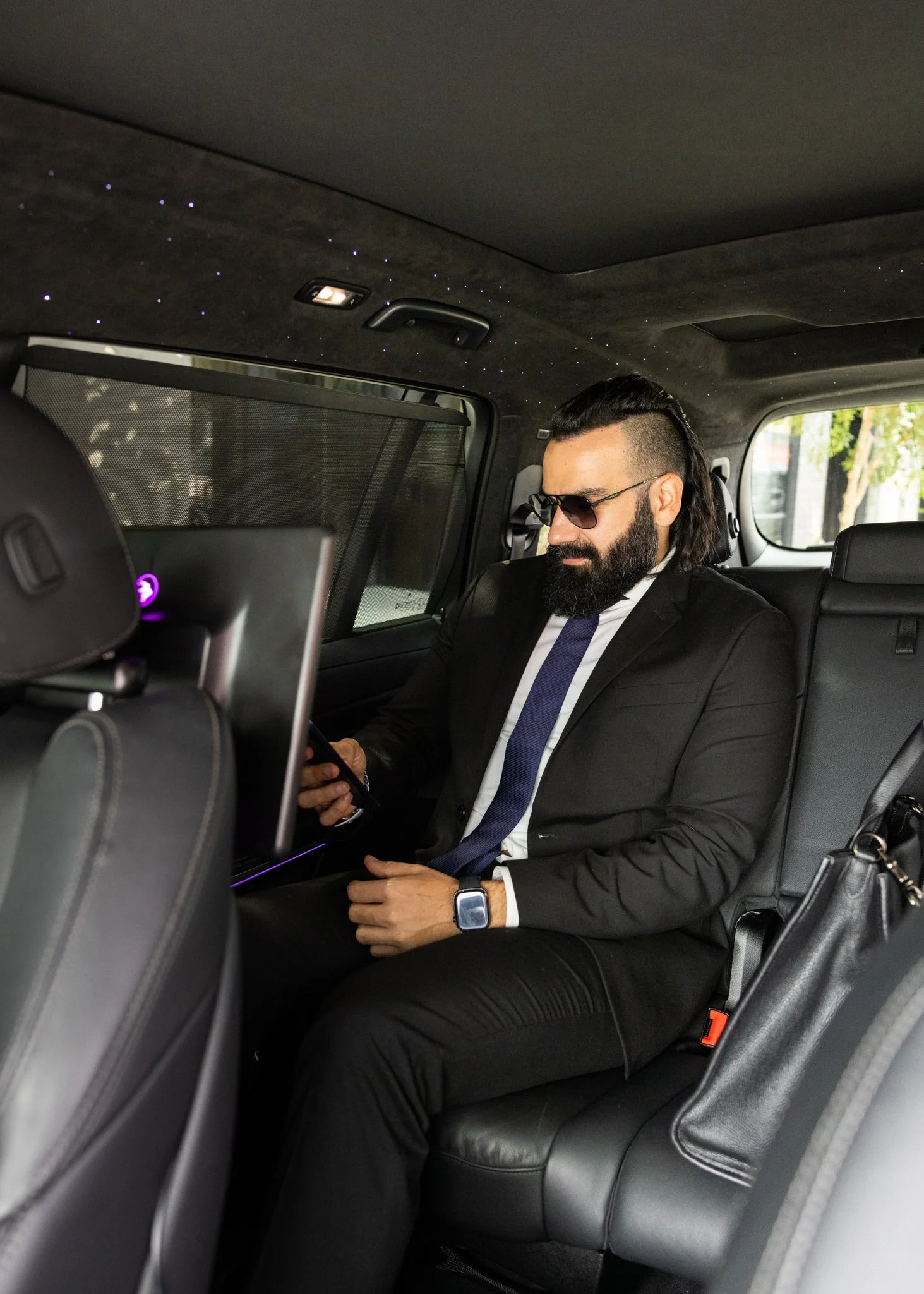 Man in a black suit, tie, sunglasses, and smartwatch sitting in the back of a car, looking at his phone.