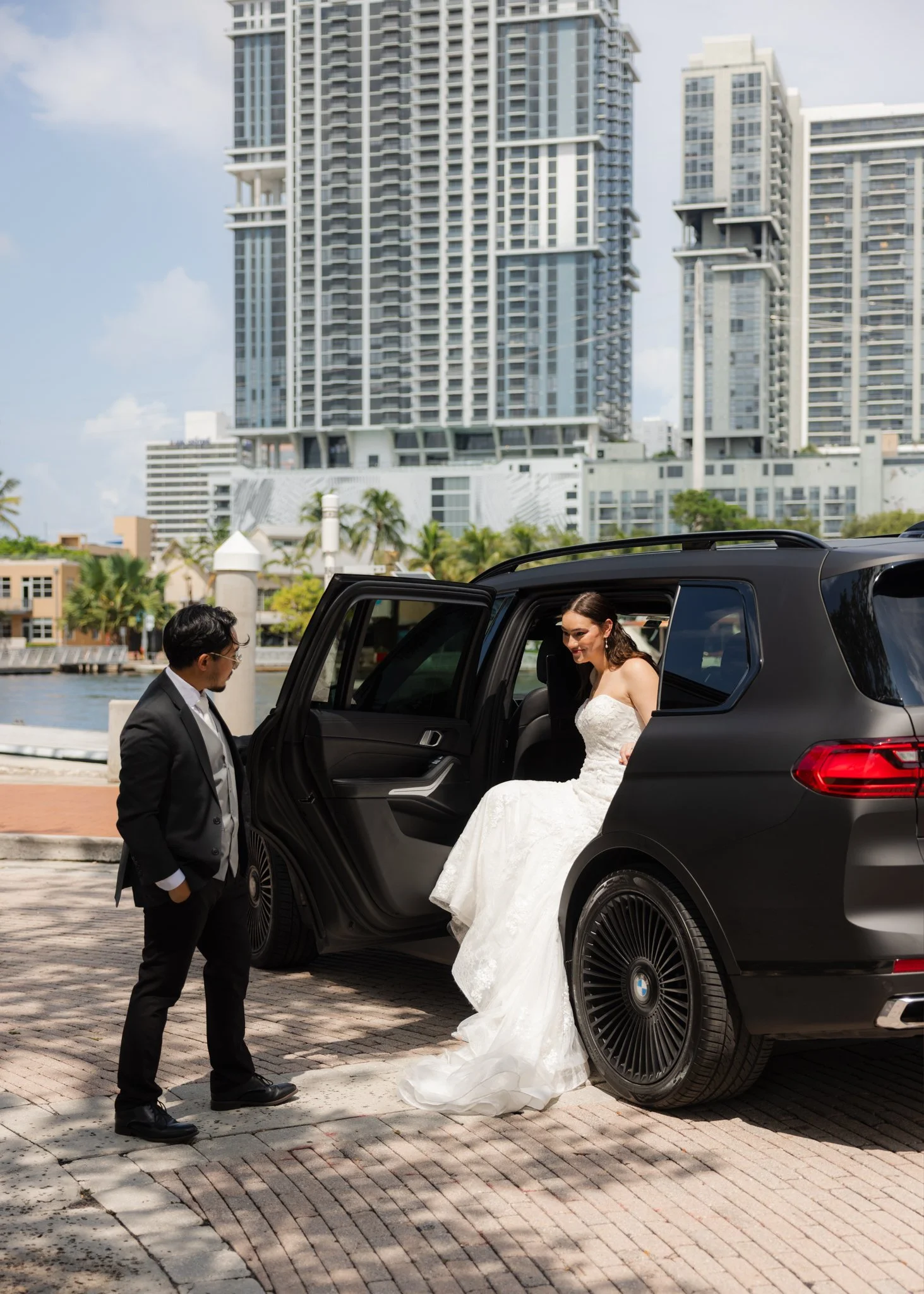 Bride in a wedding dress exiting a black SUV, with a groom in a suit standing nearby, in an urban setting with tall buildings in the background.