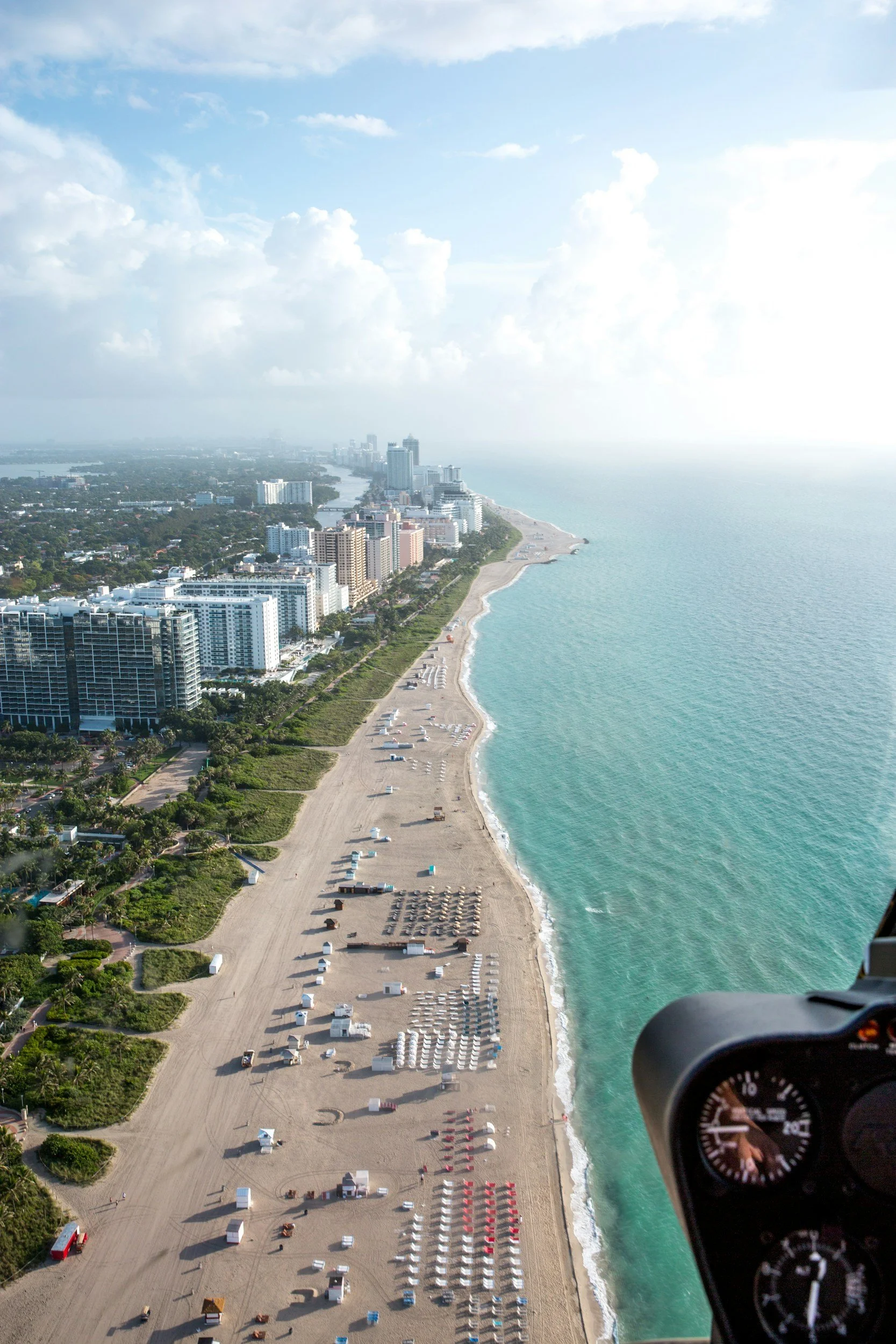 Aerial view of a beach with rows of lounge chairs, umbrellas, and small structures next to the ocean, with high-rise buildings and a city skyline in the background under a partly cloudy sky.