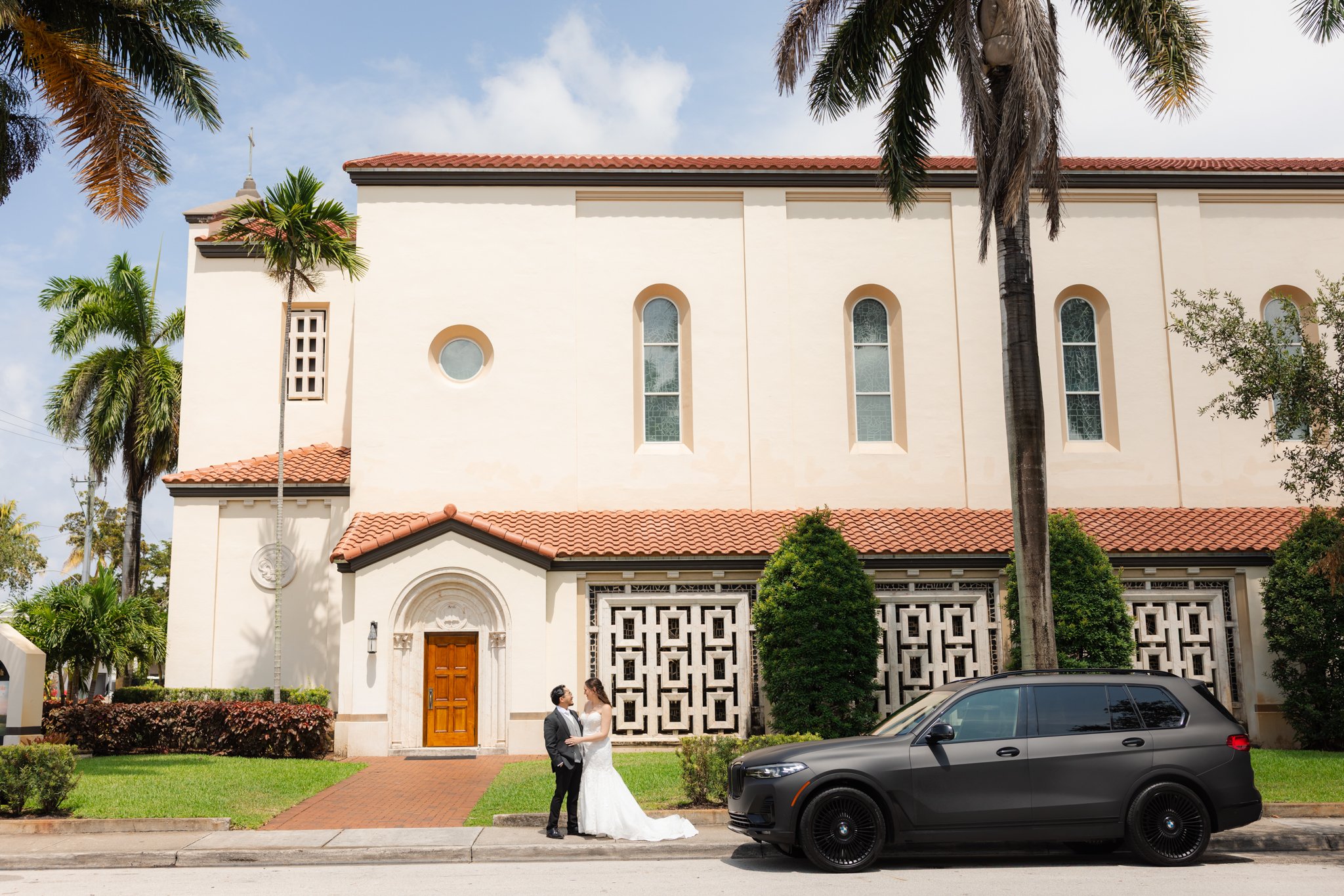 A wedding couple standing in front of a large white church with a red tile roof, palm trees, and a car parked on the street.