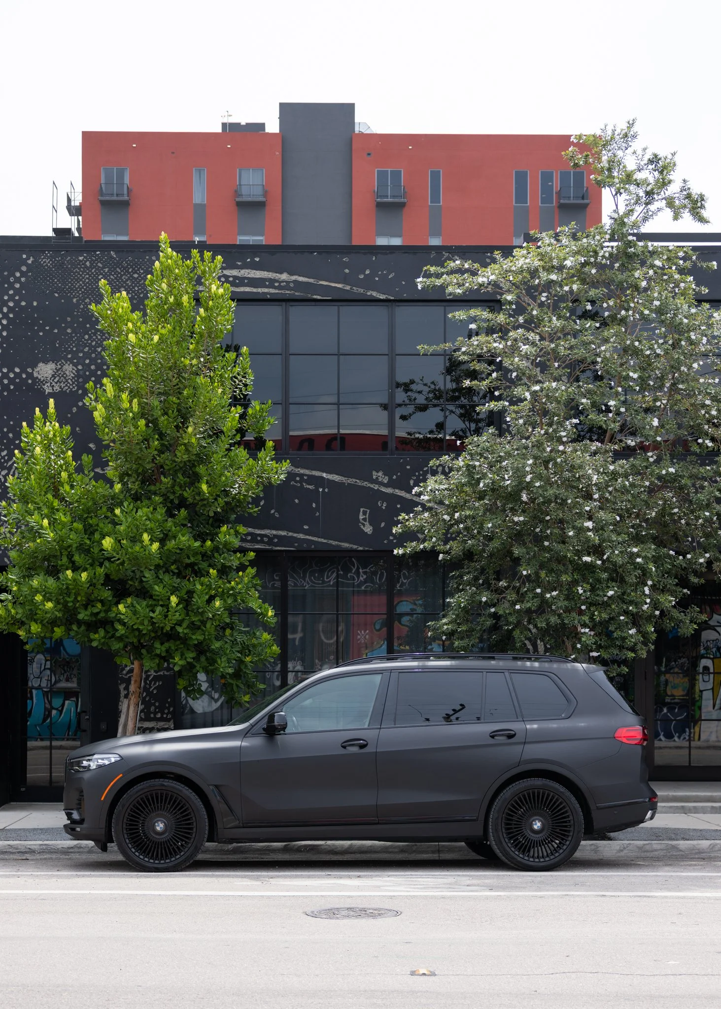 A black SUV parked on the street in front of a modern building with large windows, trees, and graffiti on the storefront. The building has a red and black façade with small balconies on upper floors.