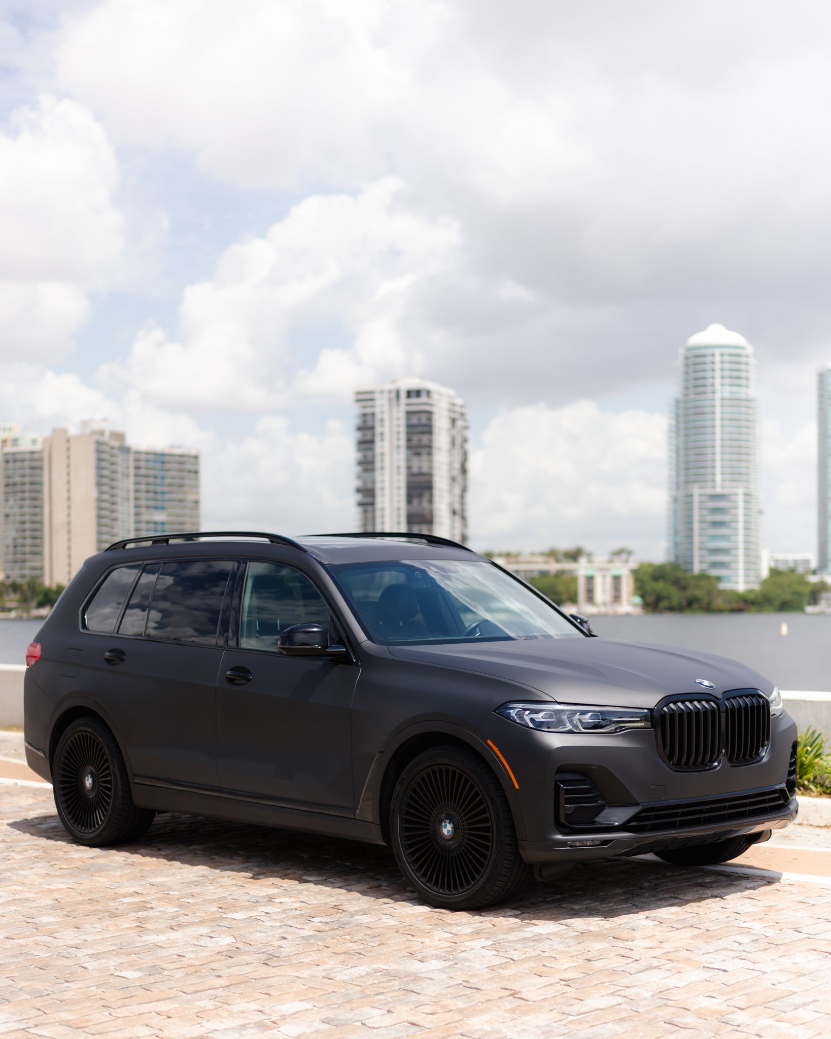 A black luxury SUV with tinted windows, black alloy wheels, and a BMW logo on the front, parked on a cobblestone surface with a city skyline of modern high-rise buildings in the background under partly cloudy skies.