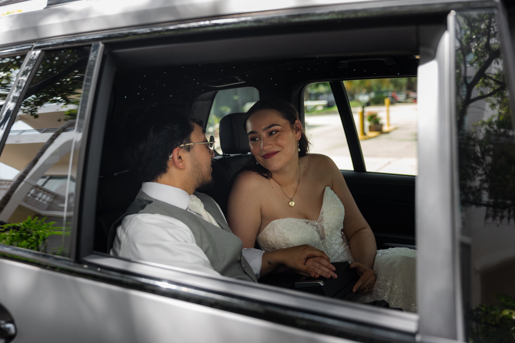 A bride and groom sitting inside a luxury vehicle, holding hands and looking at each other, with the bride wearing a wedding dress and the groom in a vest and tie, outdoors with trees and buildings in the background.