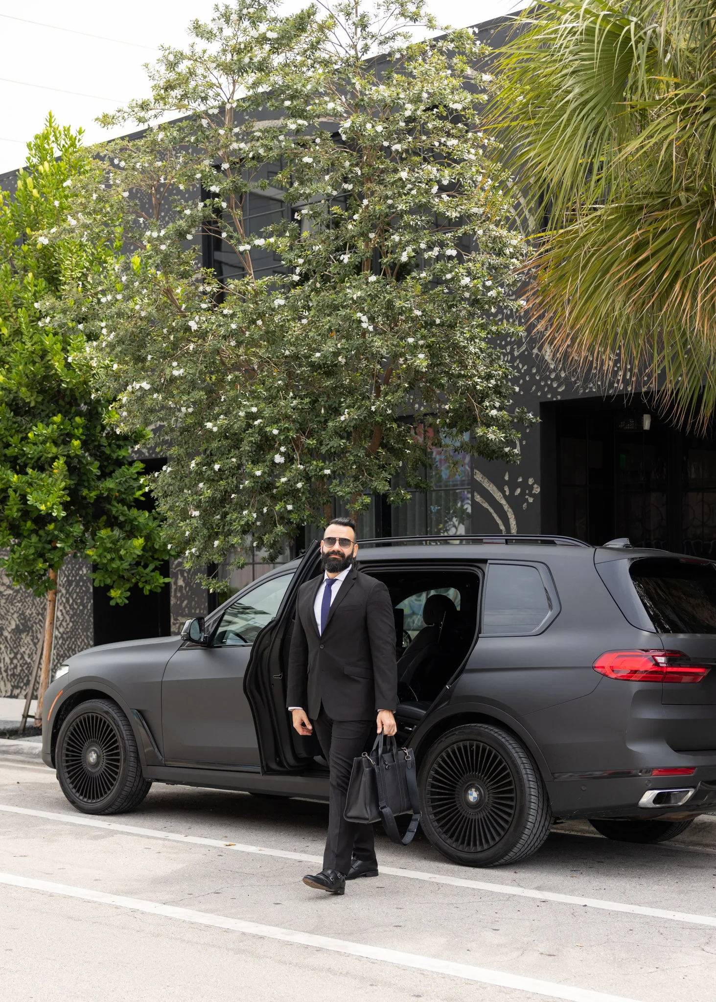 A man in a black suit, white shirt, and sunglasses standing next to a black BMW SUV with the driver's door open on a city street. He is holding a black handbag in his right hand and a black bag in his left. There are green trees and a modern building in the background.