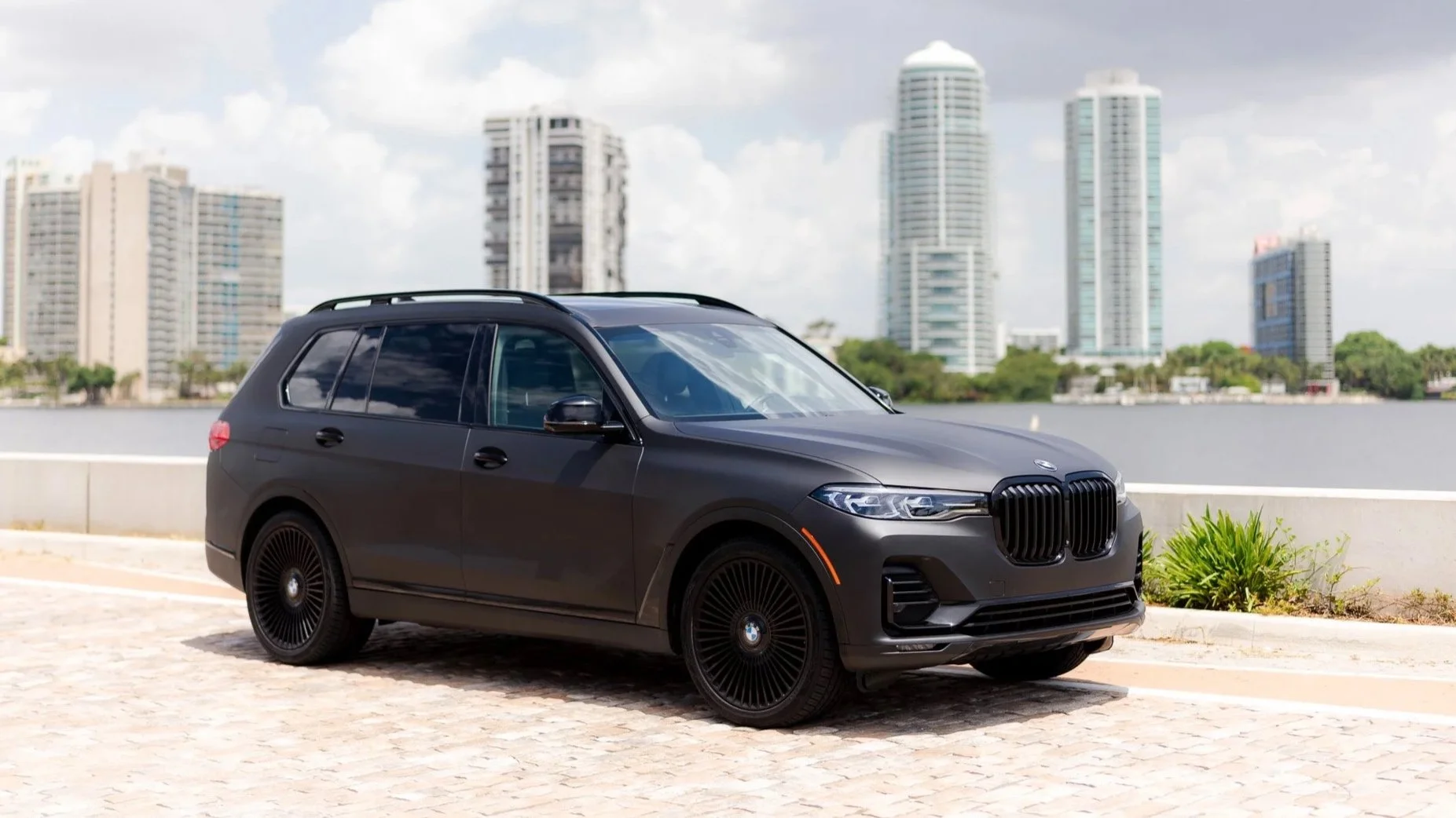 A black BMW SUV parked near a body of water with high-rise buildings in the background.