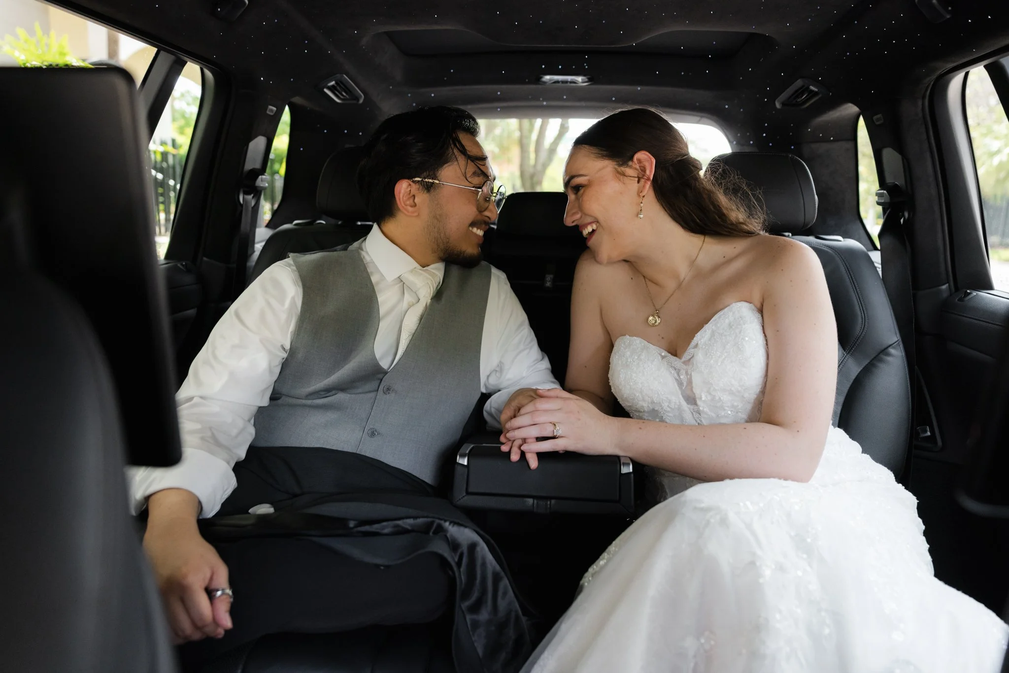 A smiling couple in wedding attire sitting in the back of a vehicle, holding hands and leaning their foreheads together.