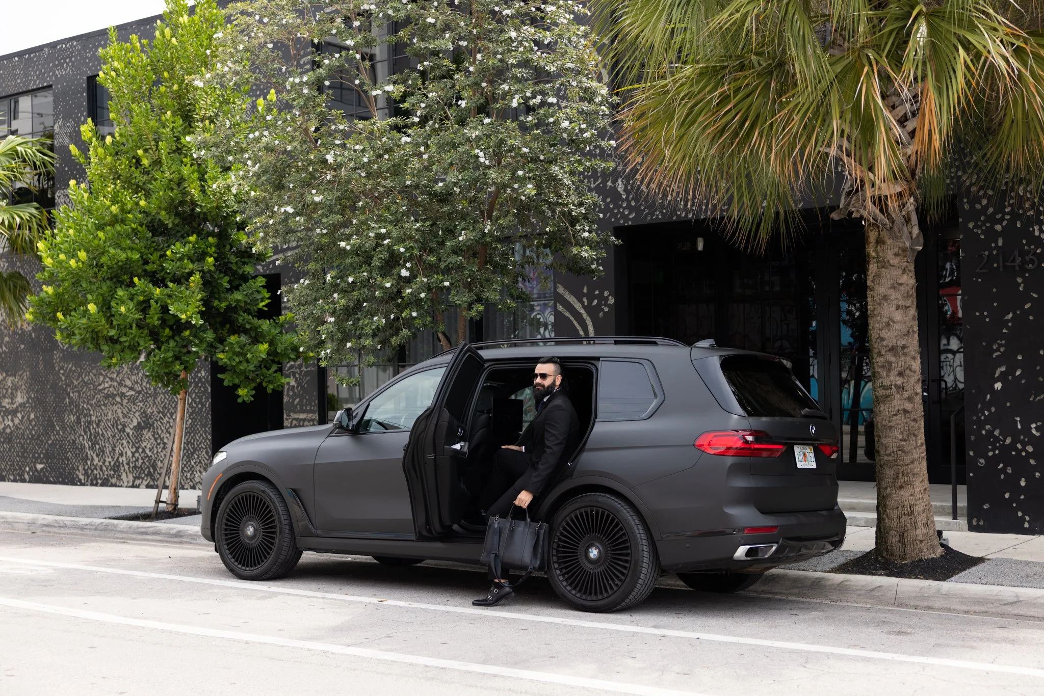 A man in a suit and sunglasses sitting in the driver's seat of a black SUV with the door open, holding a black bag, parked on a city street in front of a modern building with trees.