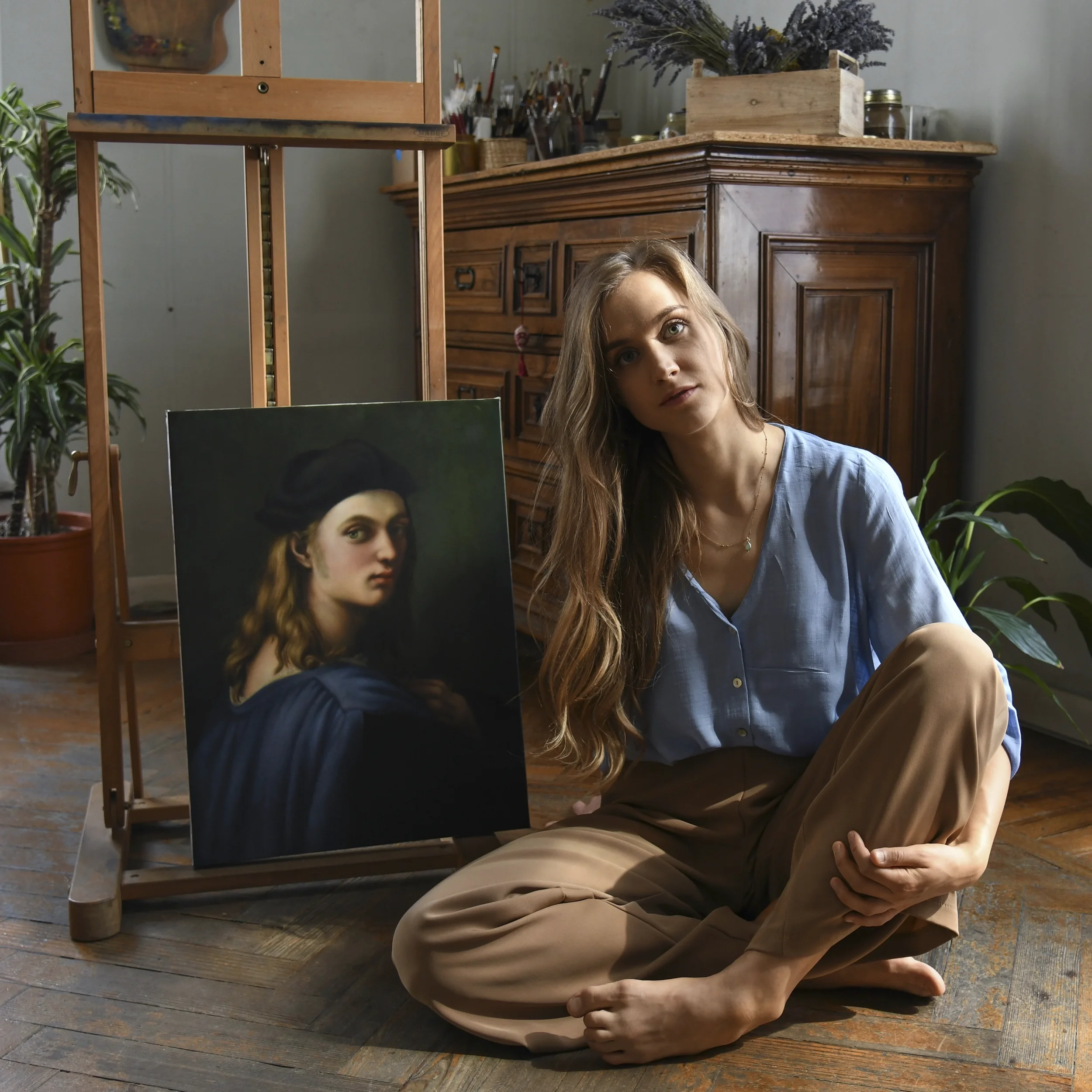 Anna Sumislawska is sitting on a wooden studio floor next to her oil painting copy of Portrait of Bindo Altoviti after Raphael. The scene shows her art studio with an easel, canvas, plants, and painting materials. 