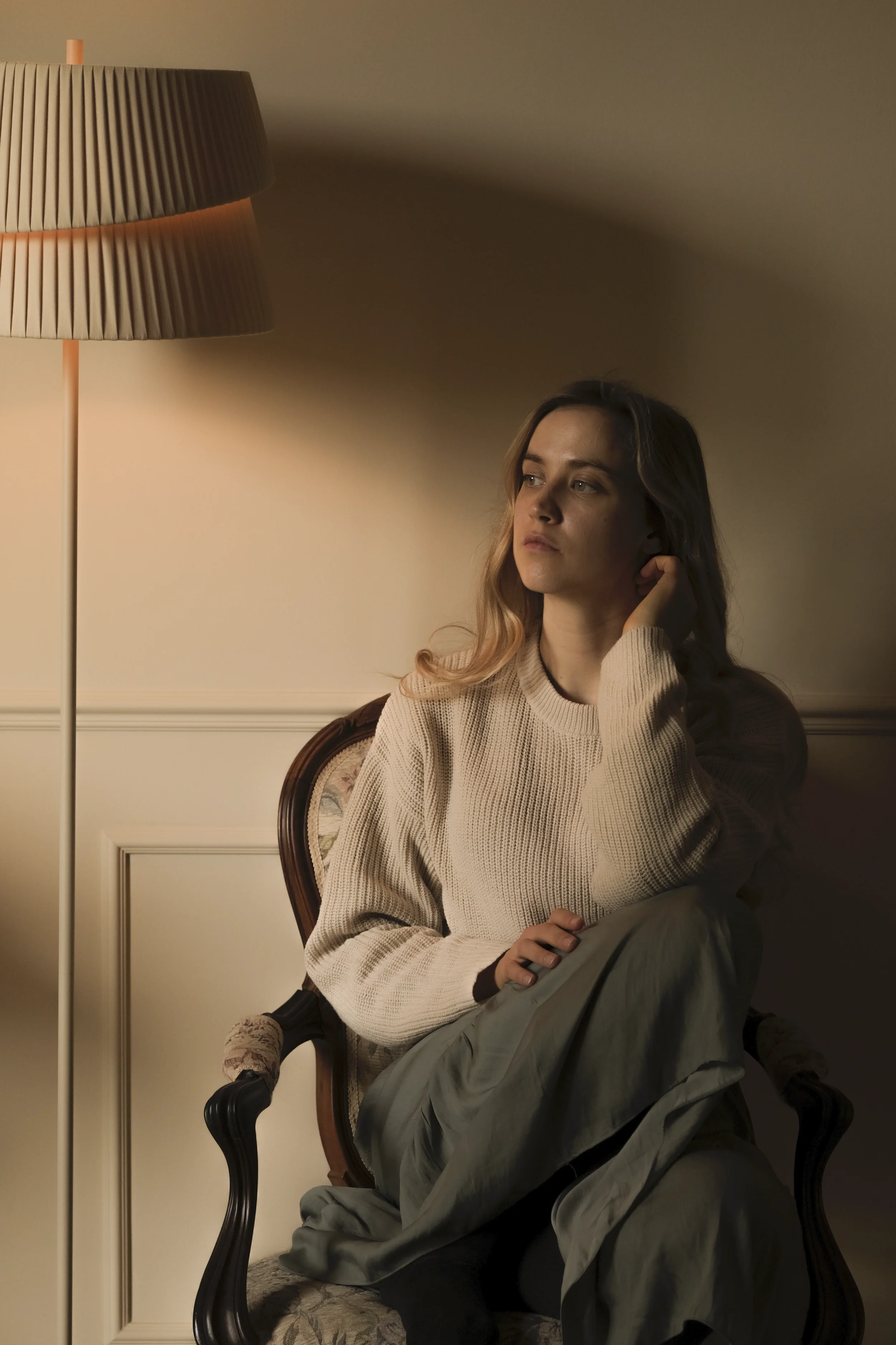 Artist Anna Sumislawska sitting in a vintage wooden armchair in a dimly lit room at home, wearing a white sweater and a muted green long dress. A tall floor lamp with a pleated shade casts warm light and soft shadows on the wall.