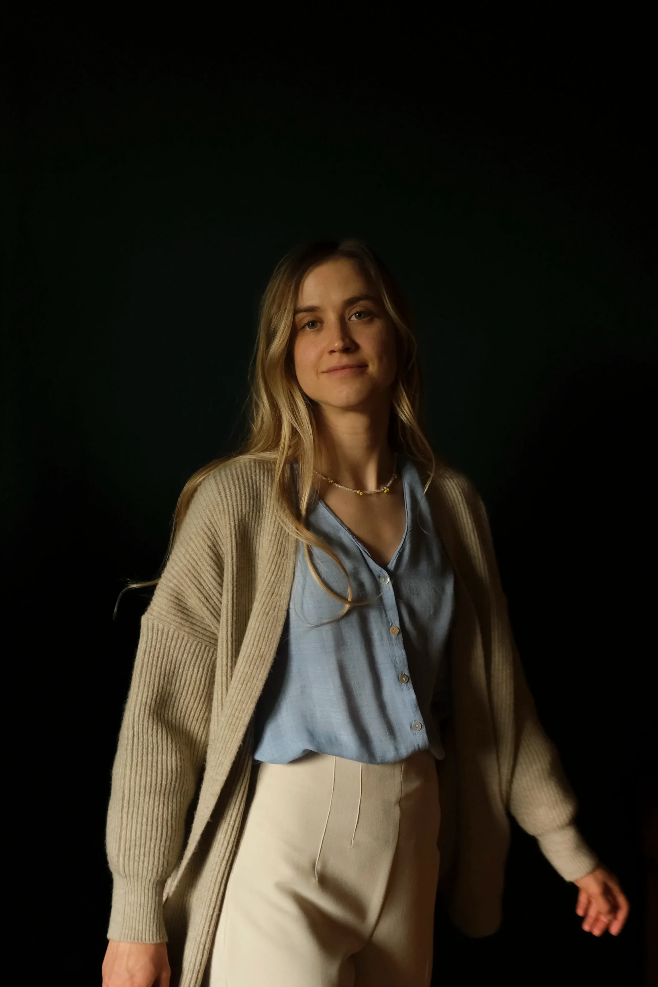 Portrait of artist Anna Sumislawska, standing against a dark background. She has long wavy blonde hair and wears a beige cardigan over a light blue blouse with cream-coloured trousers.