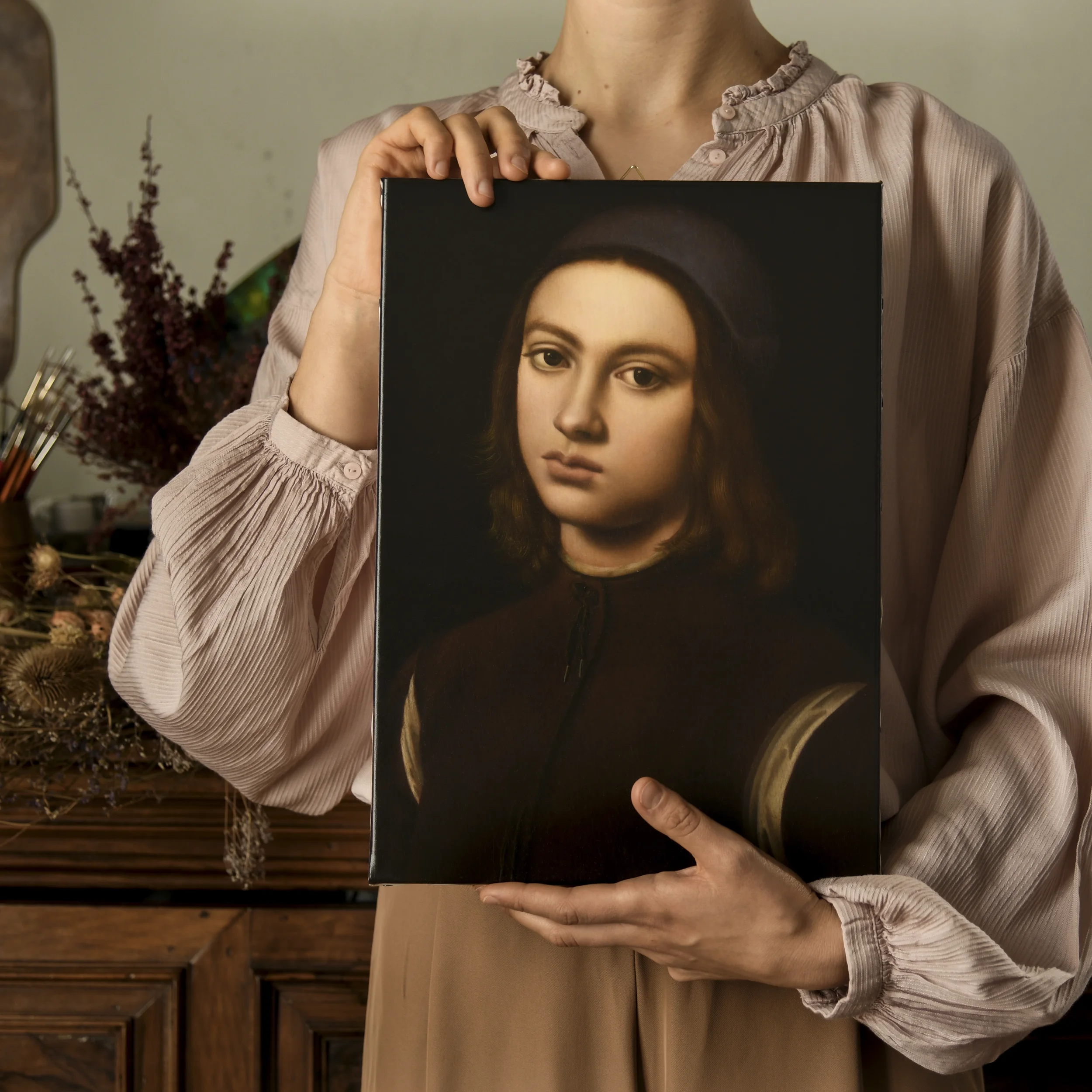 Anna Sumislawska holding a oil painting copy of a portrait of a young boy by Perugino, standing in a room with wooden furniture and dried flowers, wearing a Renaissance-style pink blouse with wide sleeves.