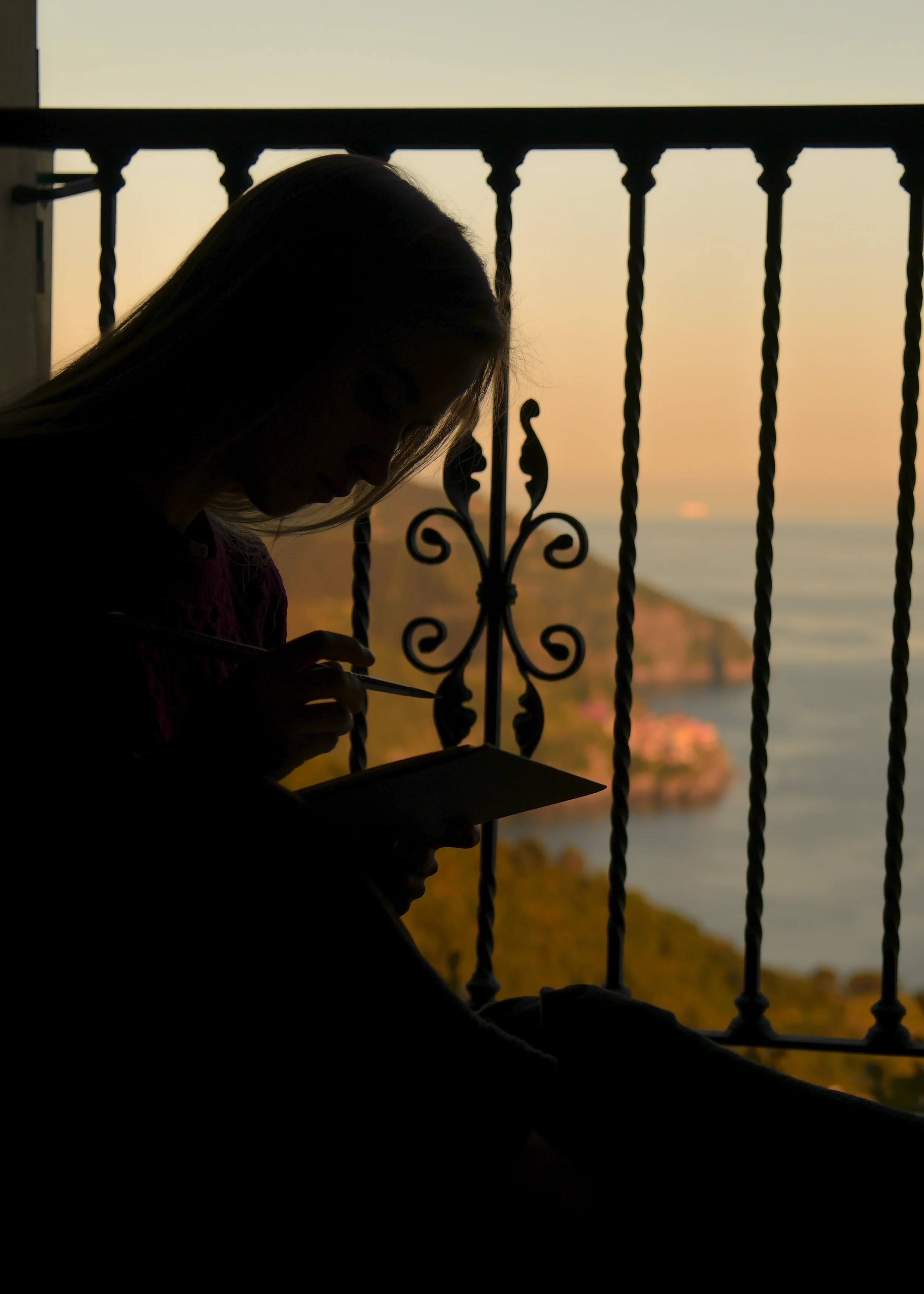 Artist Anna Sumislawska painting a watercolour seascape in the Cinque Terre, seated inside a room by a window with decorative iron railings. Through the window, the sea and a coastal village are visible at sunset.
