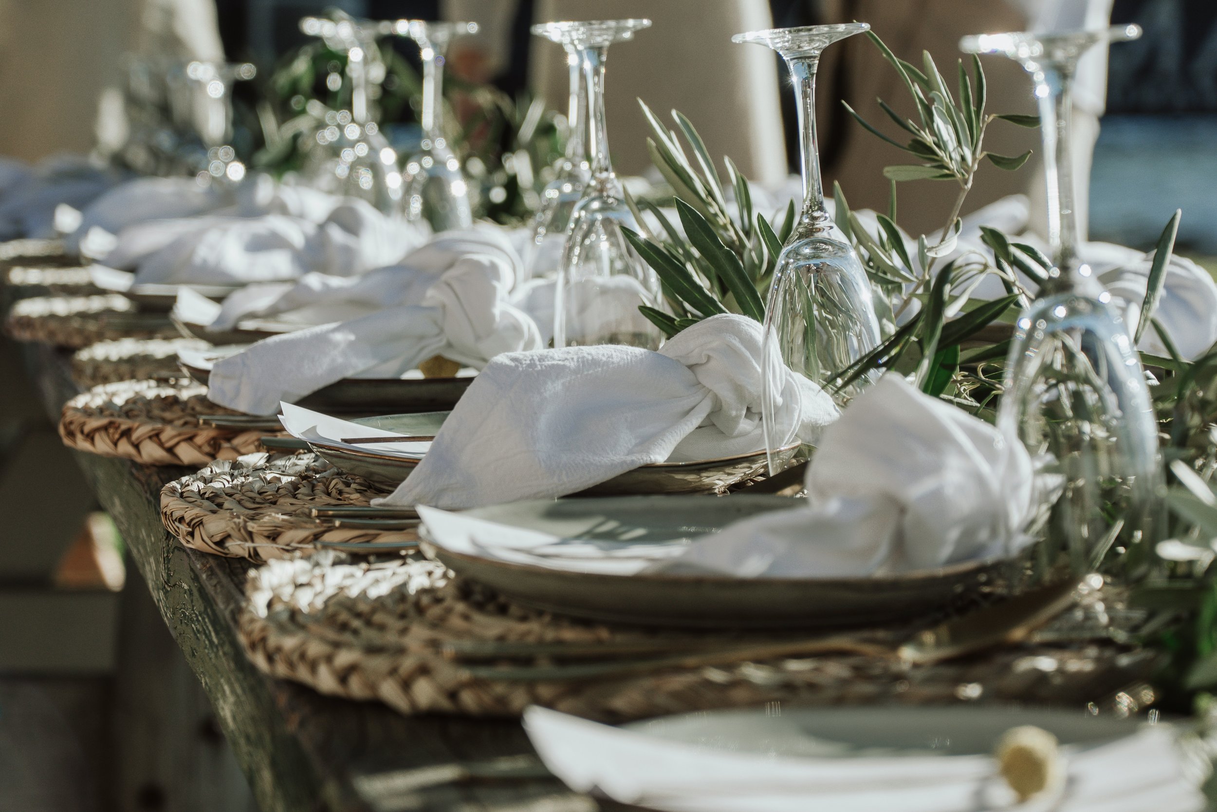 A close-up of a table set with white plates, cloth napkins tied in knots, upside-down wine glasses, woven placemats, and greenery, ready for a meal or event.