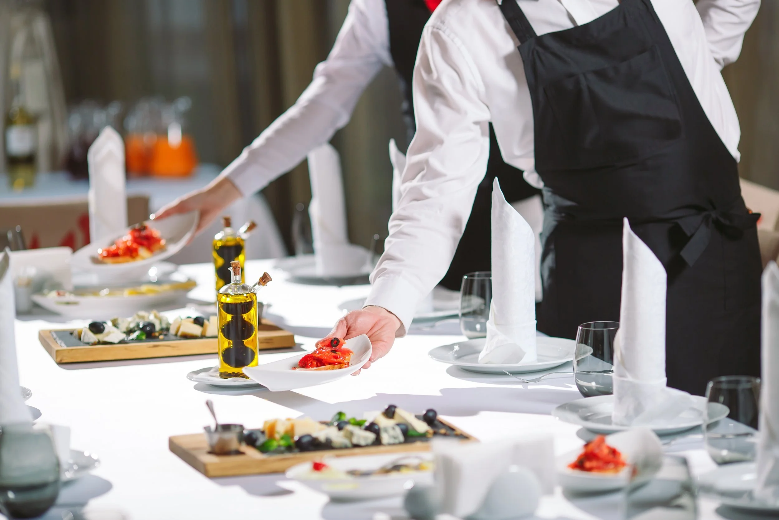 Chef in black apron serving food at a formal dining table with plates, glasses, and bottles of olive oil.