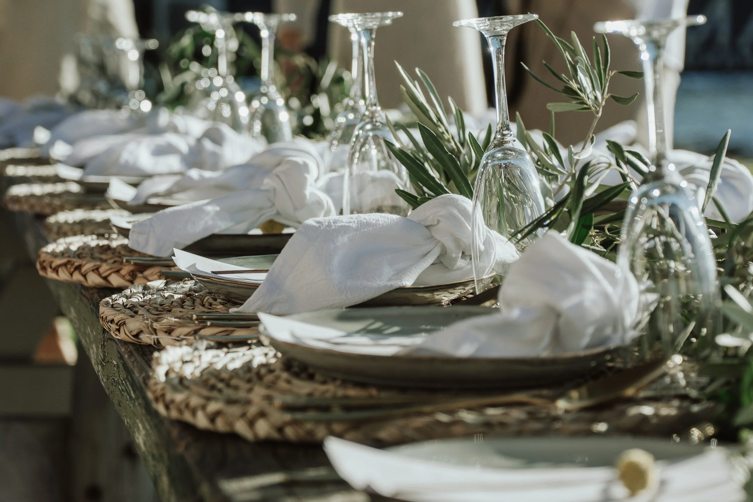 Elegant outdoor dining table set with woven placemats, white napkins tied in knots, white plates, upside-down wine glasses, and greenery decorations.