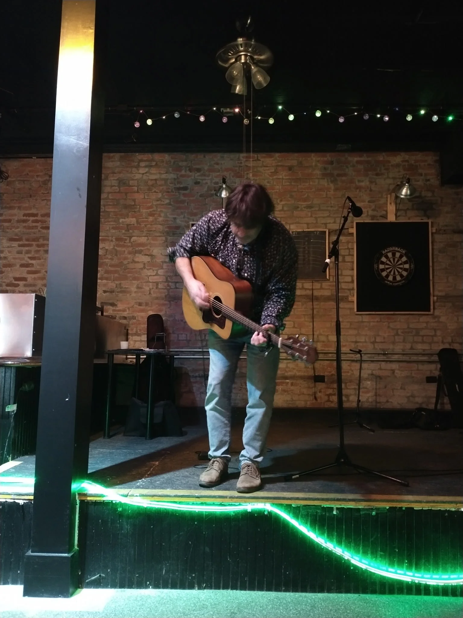 A man playing an acoustic guitar on a small stage with brick wall backdrop in a dimly lit venue decorated with colorful string lights.