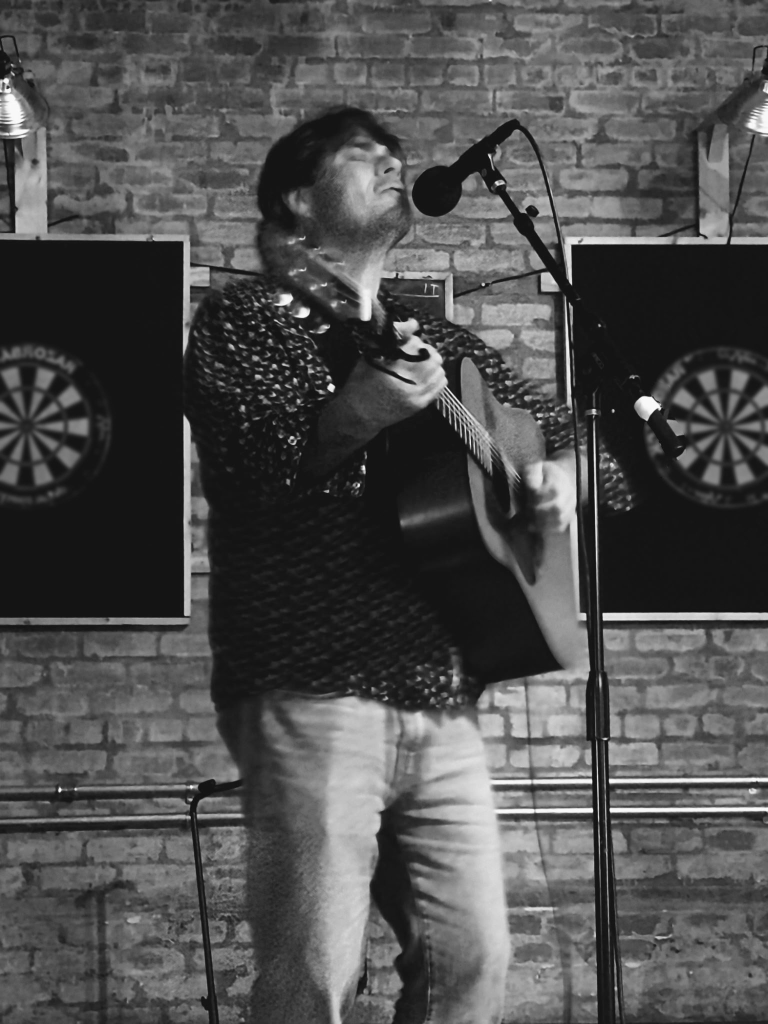 A man singing into a microphone while playing a guitar in front of a brick wall, with dartboards and two wall-mounted lights in the background.