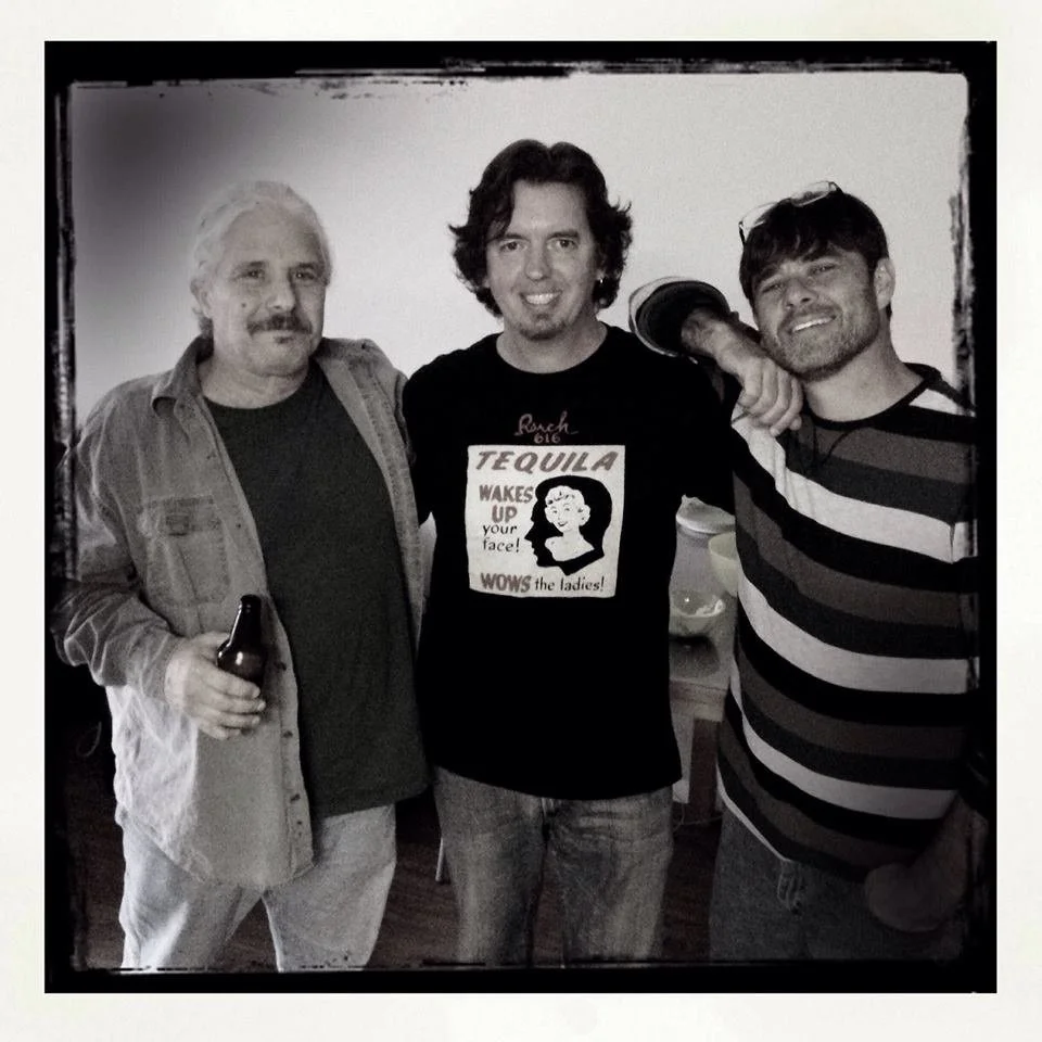 Three men standing together, smiling, in a black and white photo with a plain background.