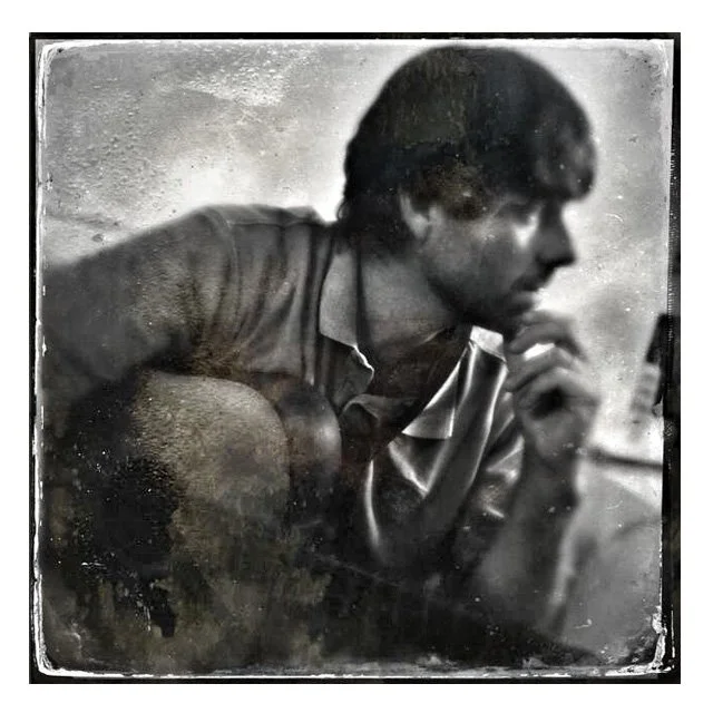 A black and white photo of a young man with dark hair, sitting with his elbow on his knee, resting his head on his hand, and holding a cigarette near his mouth, looking contemplative.