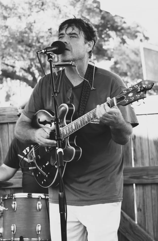 A man singing into a microphone while playing an electric guitar during an outdoor performance.