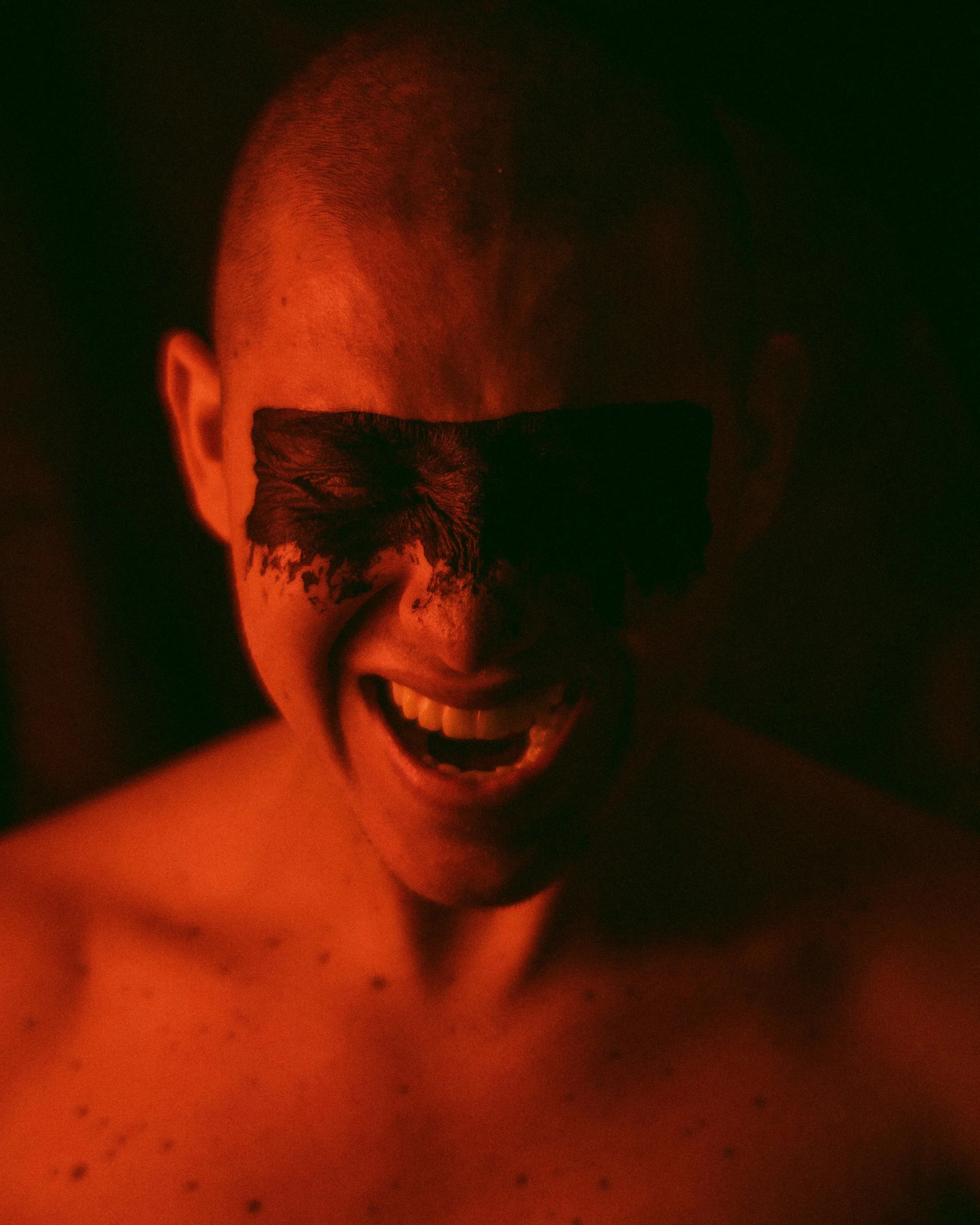 A person with a shaved head, wearing black sunglasses and smiling, illuminated by red light in a dark setting.
