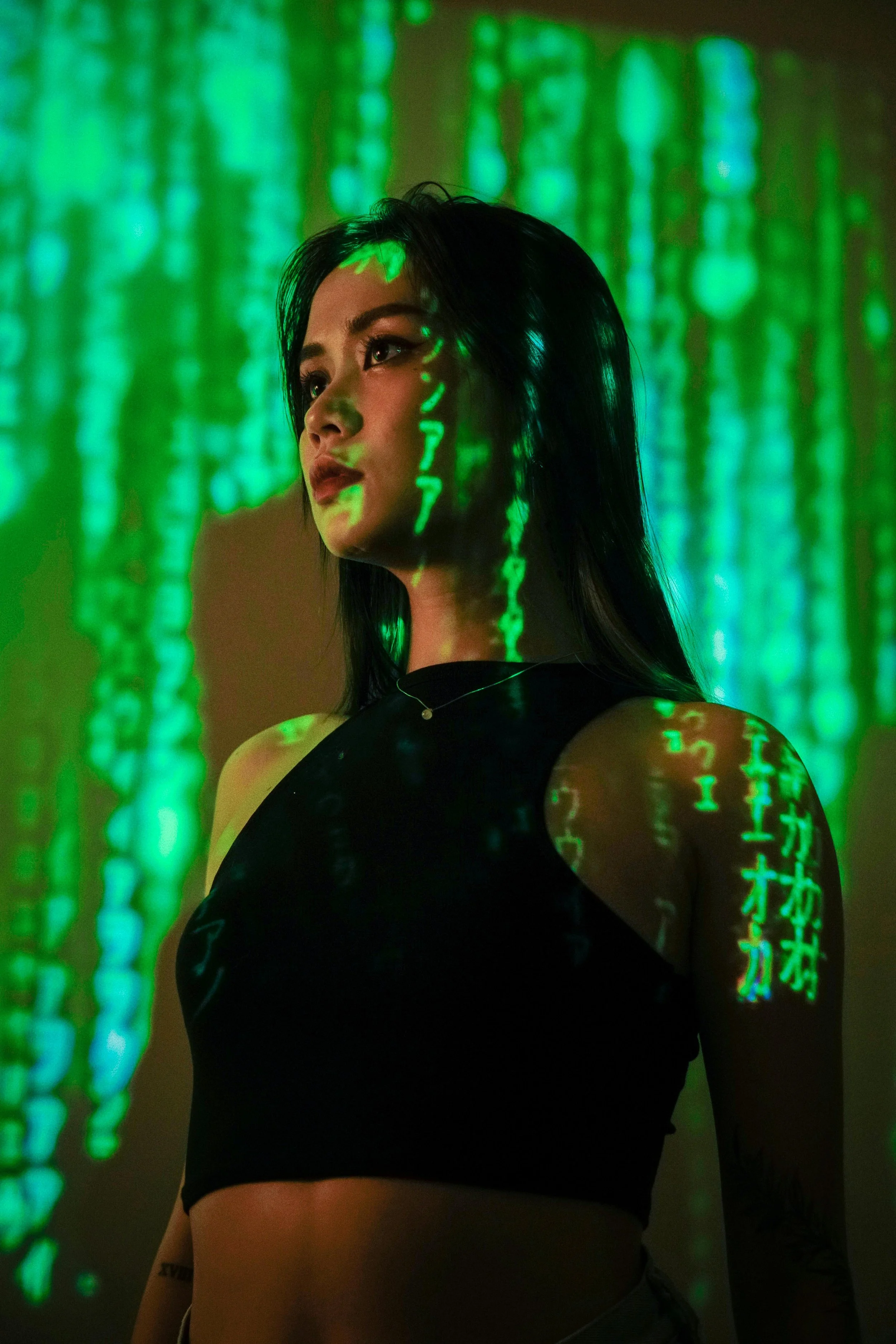 A woman with dark hair looking to the side, illuminated by green digital code projection on her face and body, with a dark background.