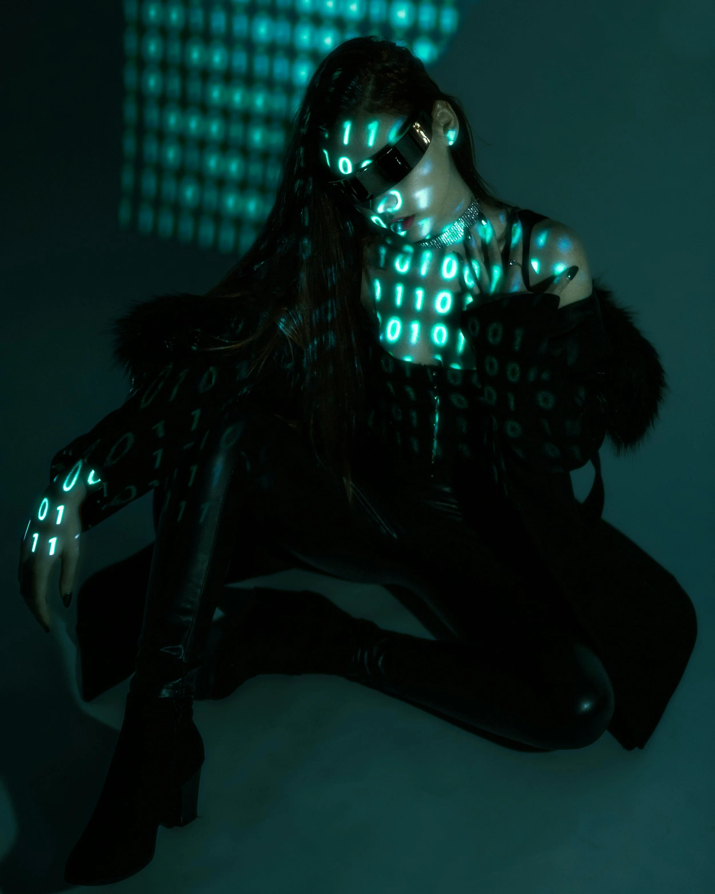 A person dressed in black with long dark hair and a choker, wearing glasses that display glowing green binary code, is sitting on a black stool against a dark background with a grid of glowing blue binary code.