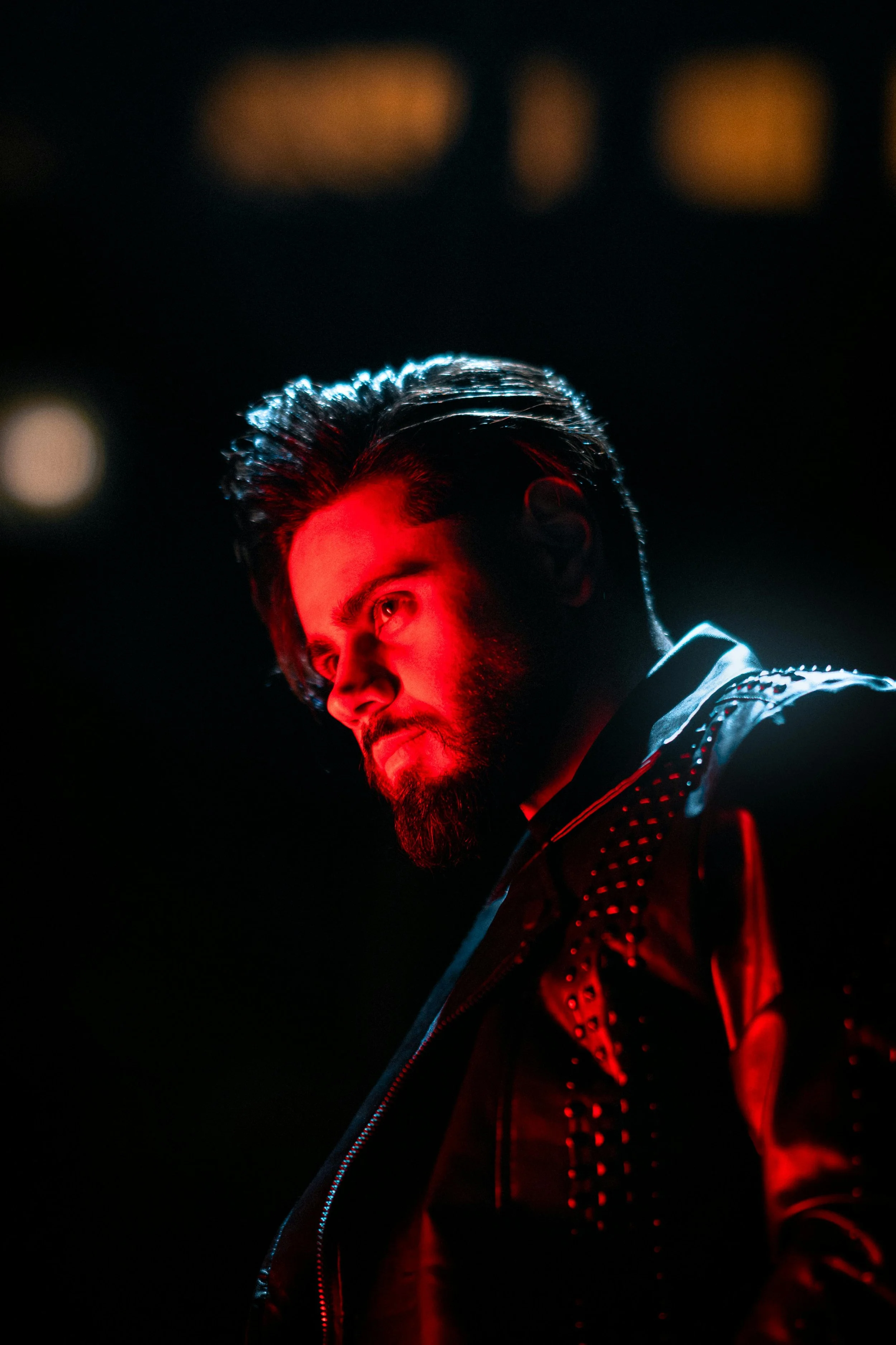 A man with dark hair and a beard, illuminated by red and blue lighting, wearing a black jacket with patterned detailing, in a dark setting.