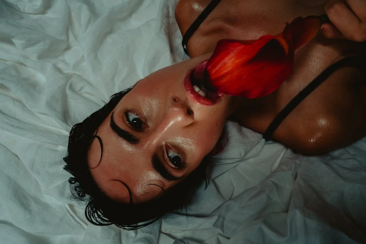 Woman on white sheets biting a red flower petal, black lingerie straps, intense close-up gaze.