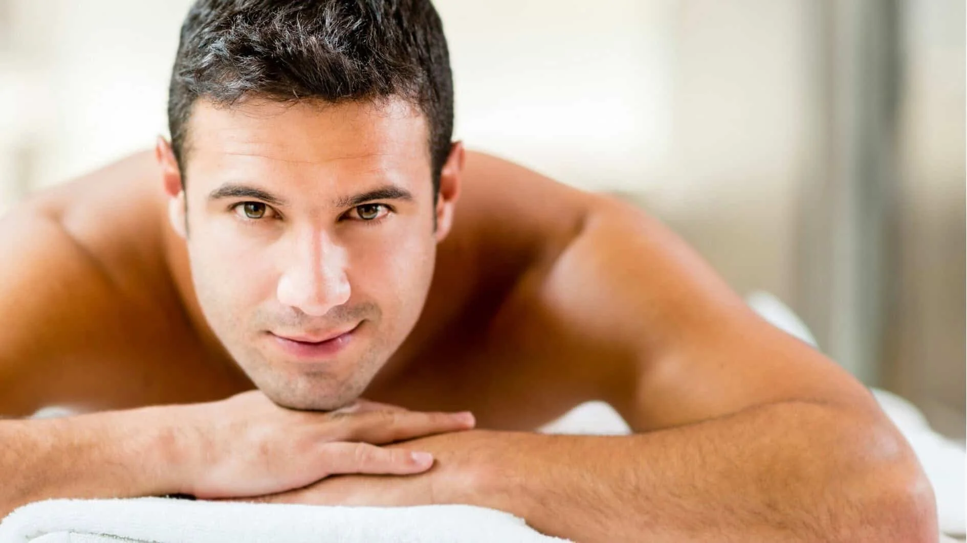 Close-up of a shirtless man lying on his stomach with his head resting on his crossed arms, looking directly at the camera with a gentle smile.