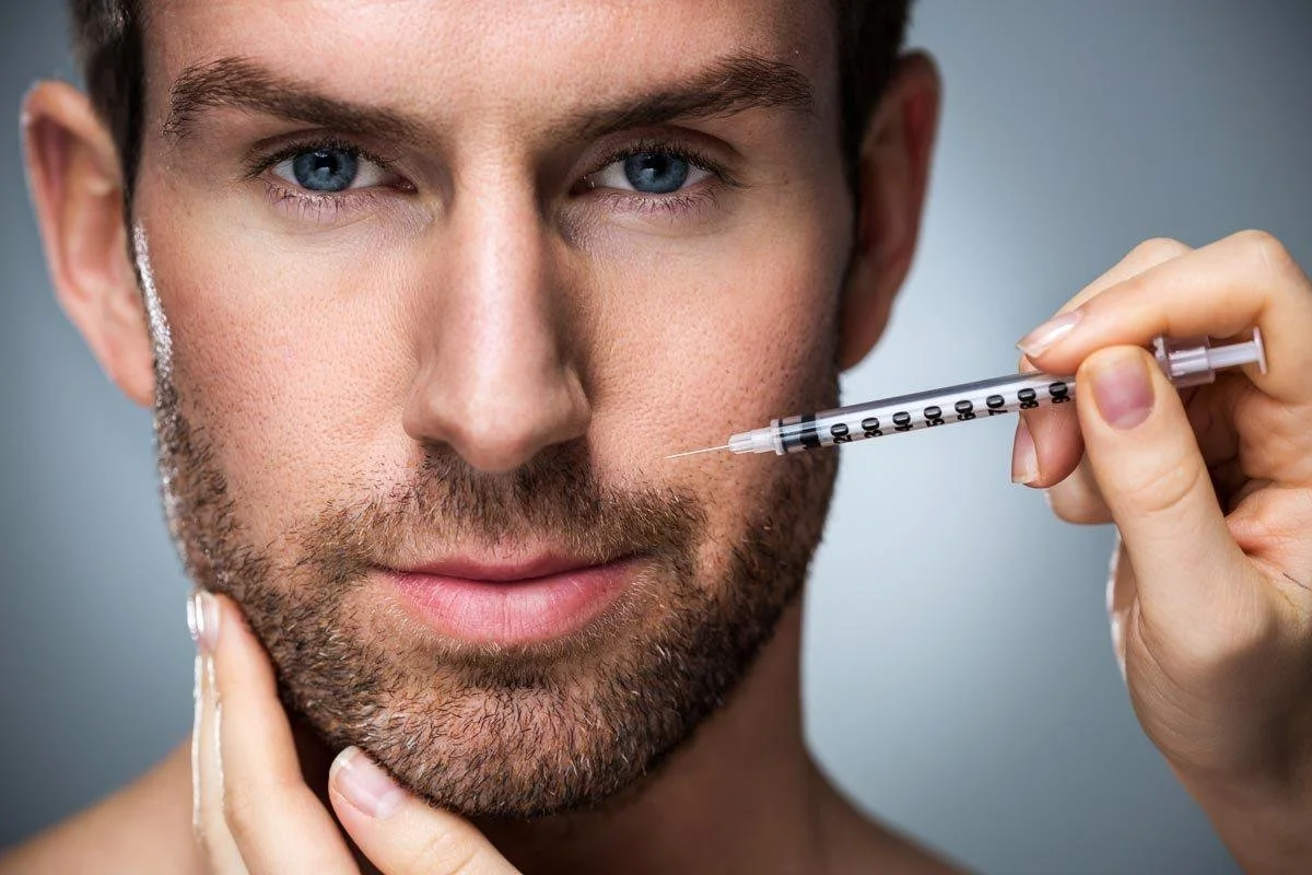 A man with blue eyes and a beard is holding a syringe near his face, looking directly at the camera.