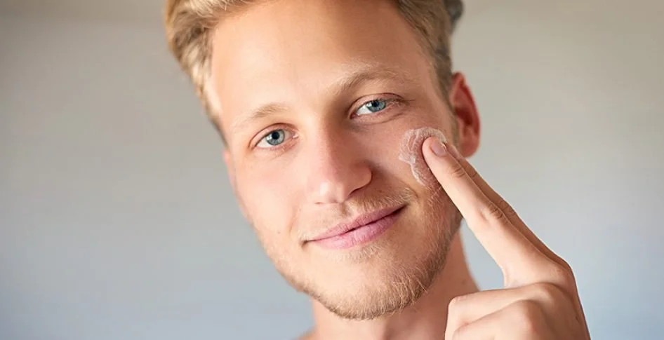 A young man with blue eyes and light hair applying facial cleanser or moisturizer to his face.