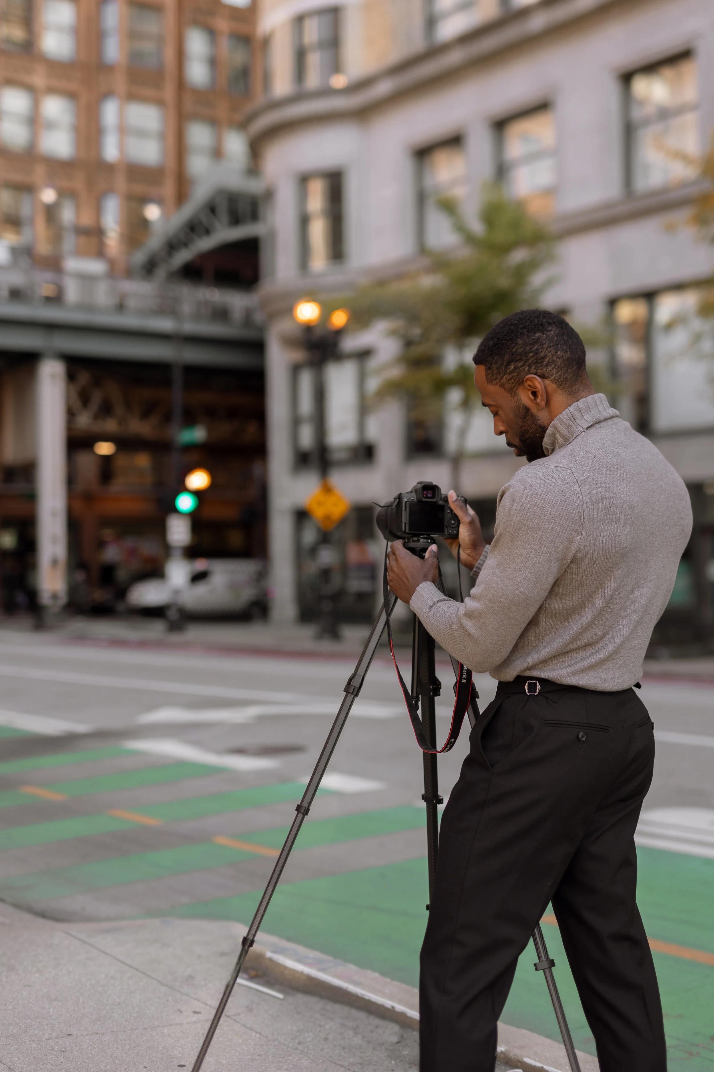 A man in business attire using a camera on a tripod outdoors in an urban setting with buildings, streetlights, and traffic signals.