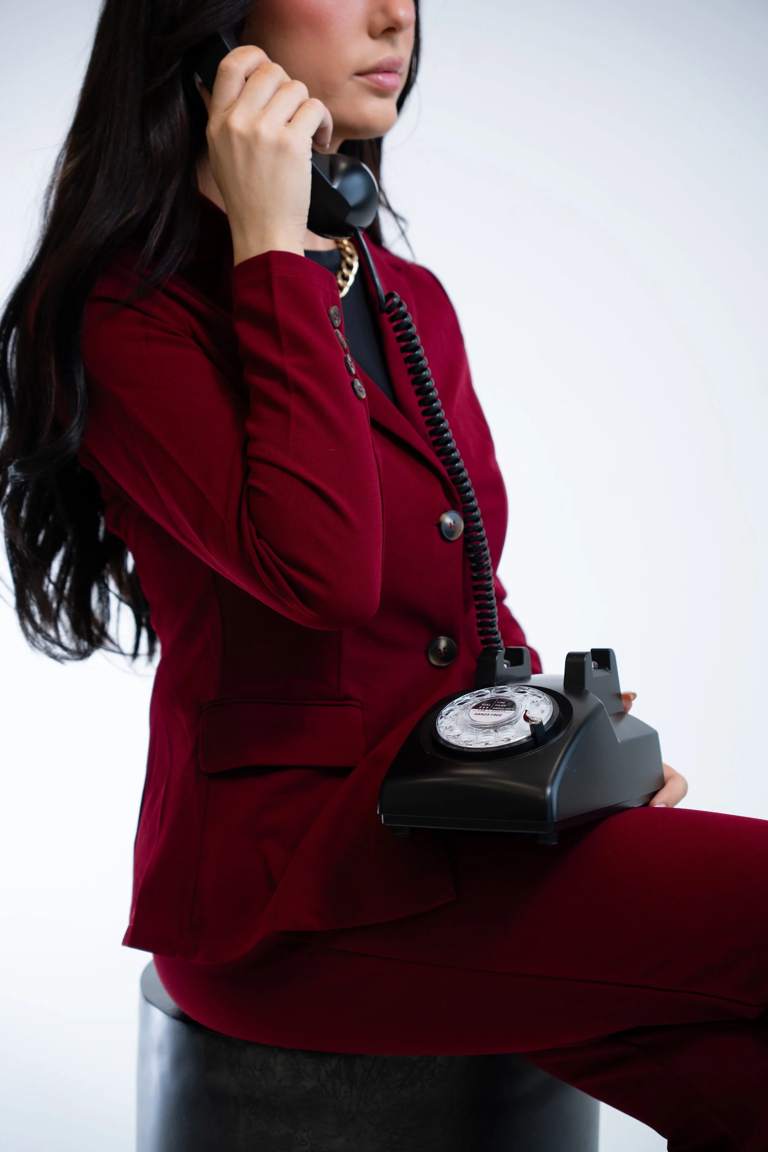 A woman dressed in a red suit sitting on a black stool, holding a vintage black rotary telephone in her hand.
