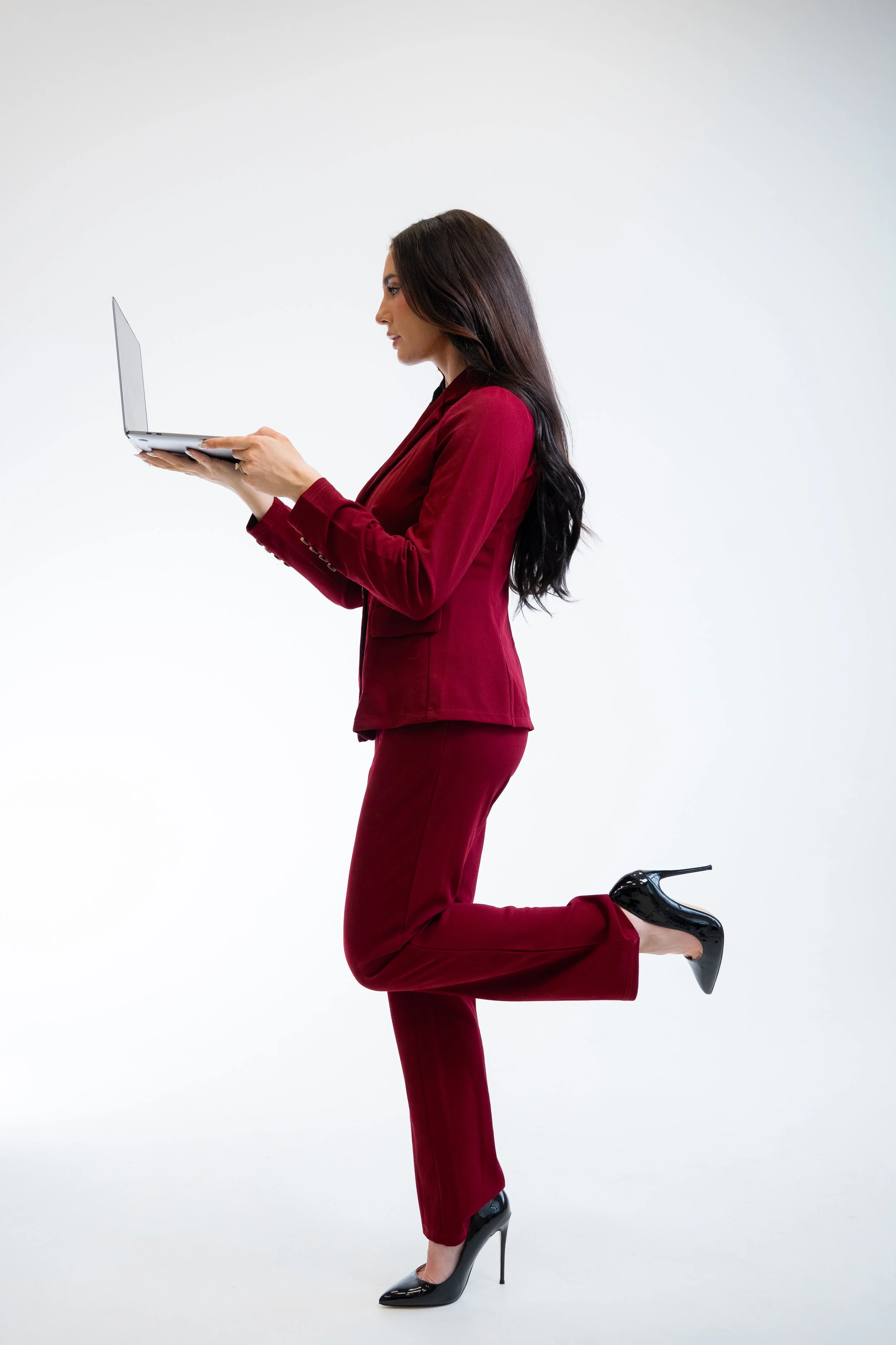 A UGC creator dressed in a red business suit with high heels is kneeling on one knee and holding a laptop in front of her against a plain white background. from a UGC Creator