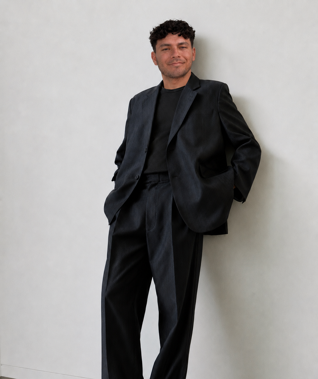 A young UGC creator in a black suit with a relaxed expression, leaning against a plain white wall, with hands in pockets.
