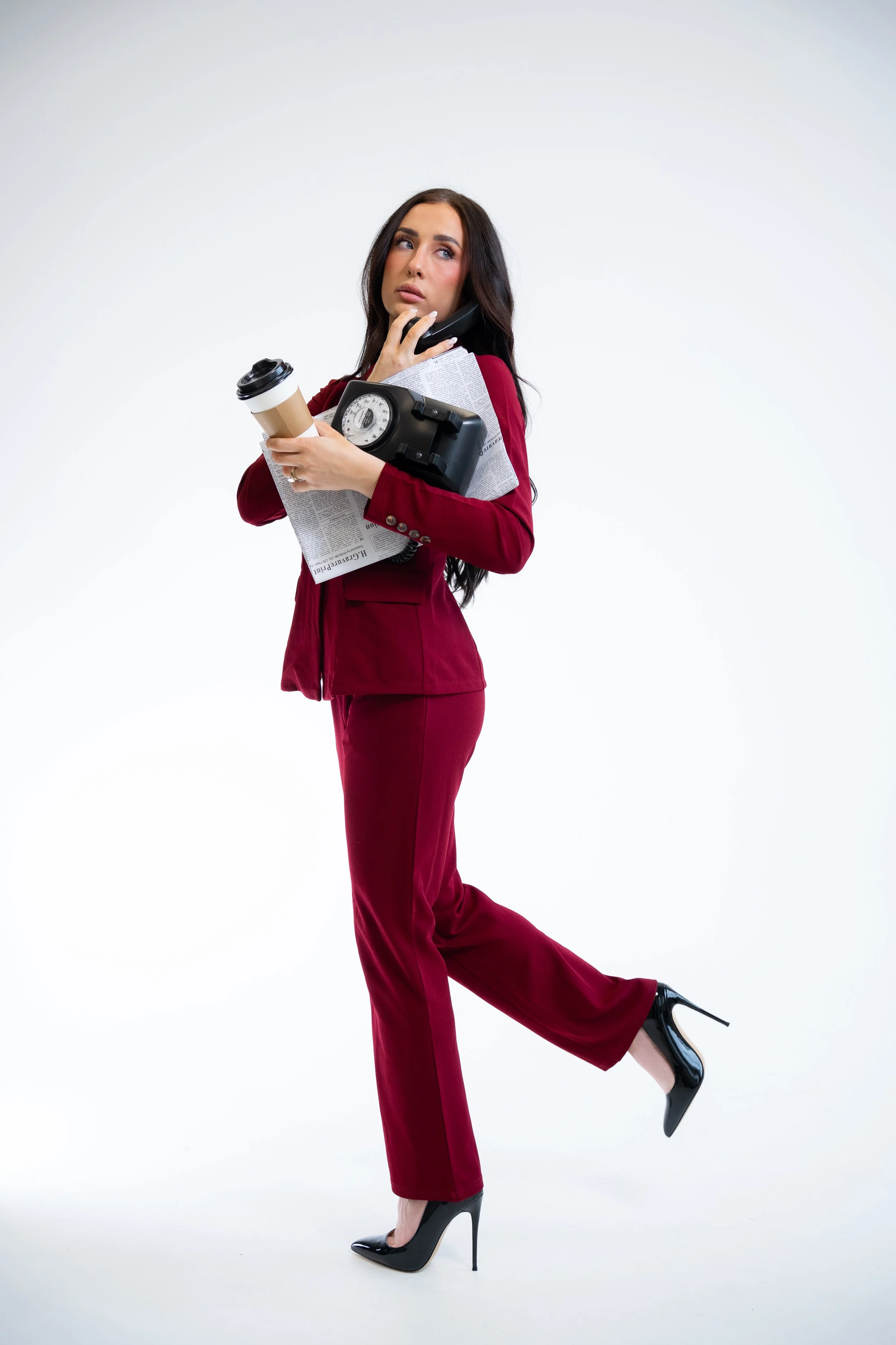 A UGC creator in a red pantsuit and black high heels holds a coffee cup, a large vintage telephone, and a newspaper, balancing on one leg with the other leg lifted behind her, against a plain white background.