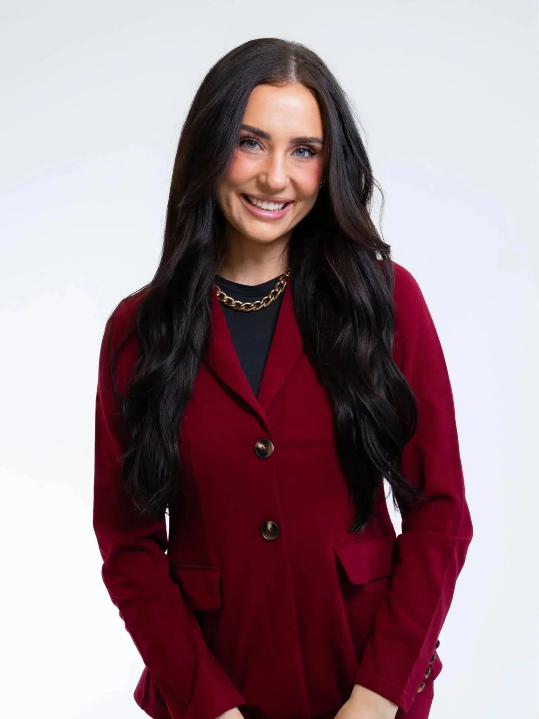 UGC creator with long dark hair, blue eyes, and a bright smile, wearing a red coat and a gold chain necklace, standing against a plain light background.