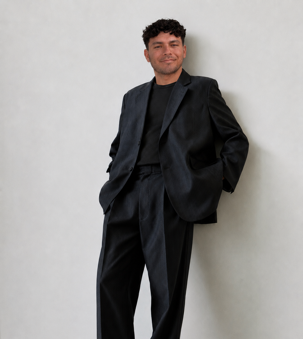 A UGC creator in a black suit with his hands in his pockets, leaning against a plain light-colored wall, smiling gently at the camera.