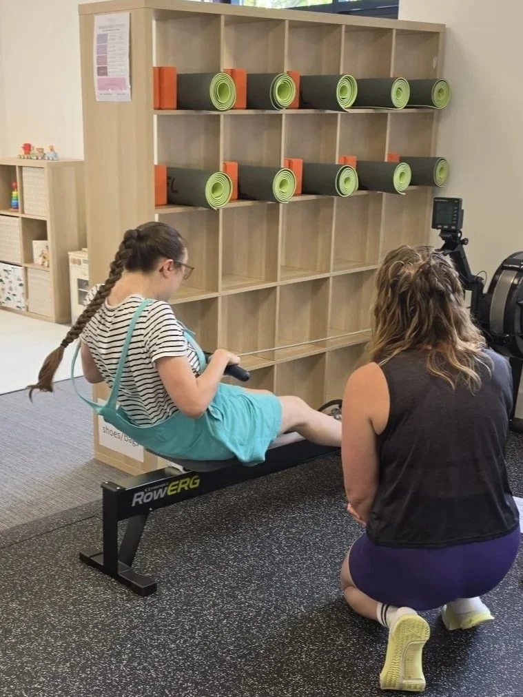 A girl with a braided hair wearing glasses, a striped shirt, and turquoise shorts, is seated on a rowing machine during a fitness assessment with a woman kneeling beside her. In the background, there is a shelving unit with green and black foam rollers and orange brackets.